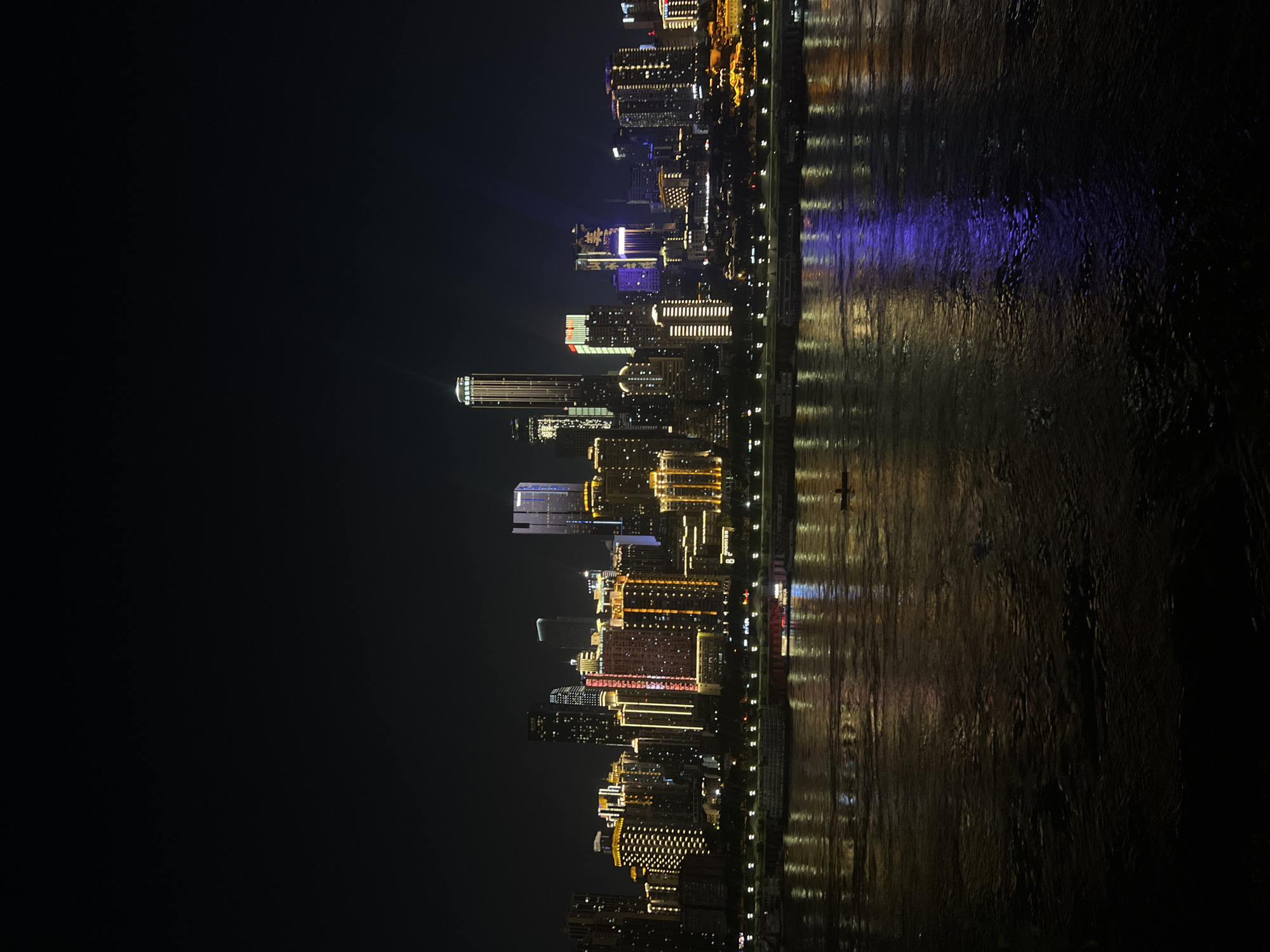 A nighttime panoramic view of the cityscape of Nan'an District, Chongqing, China, showcases a dense collection of illuminated skyscrapers and buildings along the waterfront. The structures are adorned with a variety of colorful lights, predominantly in shades of yellow, orange, white, and some blue. A wide, dark body of water, reflecting the city lights, dominates the foreground. Several boats and ferries are visible docked along the riverbank. The sky above is a deep, starless black. The scene captures a vibrant urban environment at night, with the illuminated skyline creating a striking contrast against the dark water and sky.