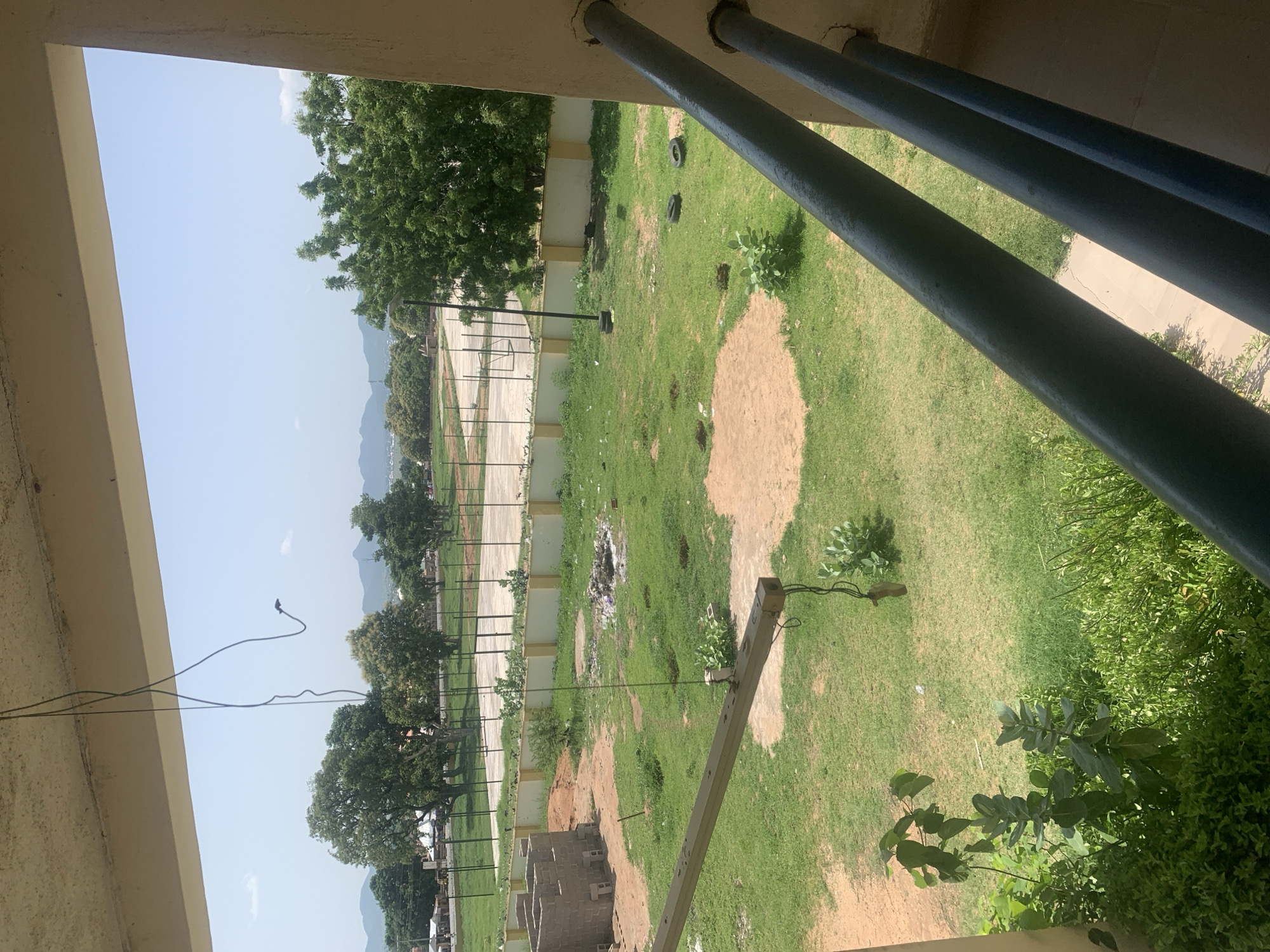 The image depicts an outdoor scene, likely a sports field or recreational area, viewed from a covered walkway. In the foreground, a beige-colored overhang and dark blue metal pipes are visible, suggesting the perspective is from a building's exterior.
The main subject is a large grassy area with patches of dirt and some scattered debris, including what appears to be trash. There are several trees of varying sizes, some with dense green foliage, located around the perimeter and within the open space. A paved area, possibly a court or pathway, runs along the left side of the frame, marked with white lines and a net or fence. In the distance, a chain-link fence encloses another section of the field. Further beyond that, a line of smaller trees and buildings can be seen, leading up to hills or mountains in the far background.
The sky is a clear, pale blue, indicating daytime with fair weather. The lighting suggests it is likely morning or late afternoon, given the angle of shadows, though this is not definitively clear. There is no clear activity or event taking place; the scene appears to be a static view of the location. No people are visible in the image.
Notable details include two car tires lying on the grass, a wooden pole with some wires attached, and what appears to be partially constructed brick walls in the lower left portion of the image. The presence of these elements suggests ongoing development or a lack of regular maintenance in parts of the area. The overall impression is of a somewhat unkempt but functional outdoor space in a warm climate, possibly in a developing area.