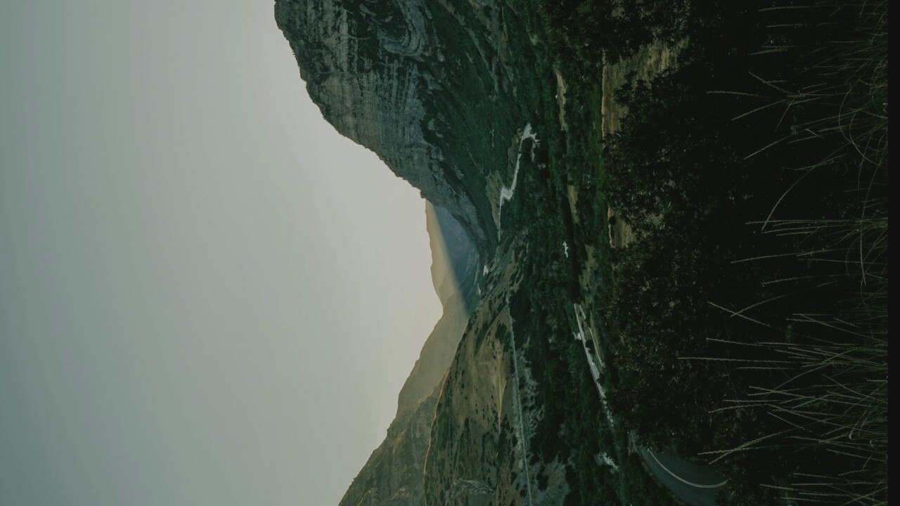 A winding road cuts through a valley carved by mountains. The sky is a pale, washed-out blue. Sunlight streams through a gap in the mountains, casting a dramatic beam across the valley floor. The slopes of the mountains are covered in sparse vegetation, with darker green trees and bushes filling the valley. In the foreground, tall, dry grass stands silhouetted against the darker foliage. The scene evokes a sense of natural grandeur and isolation.