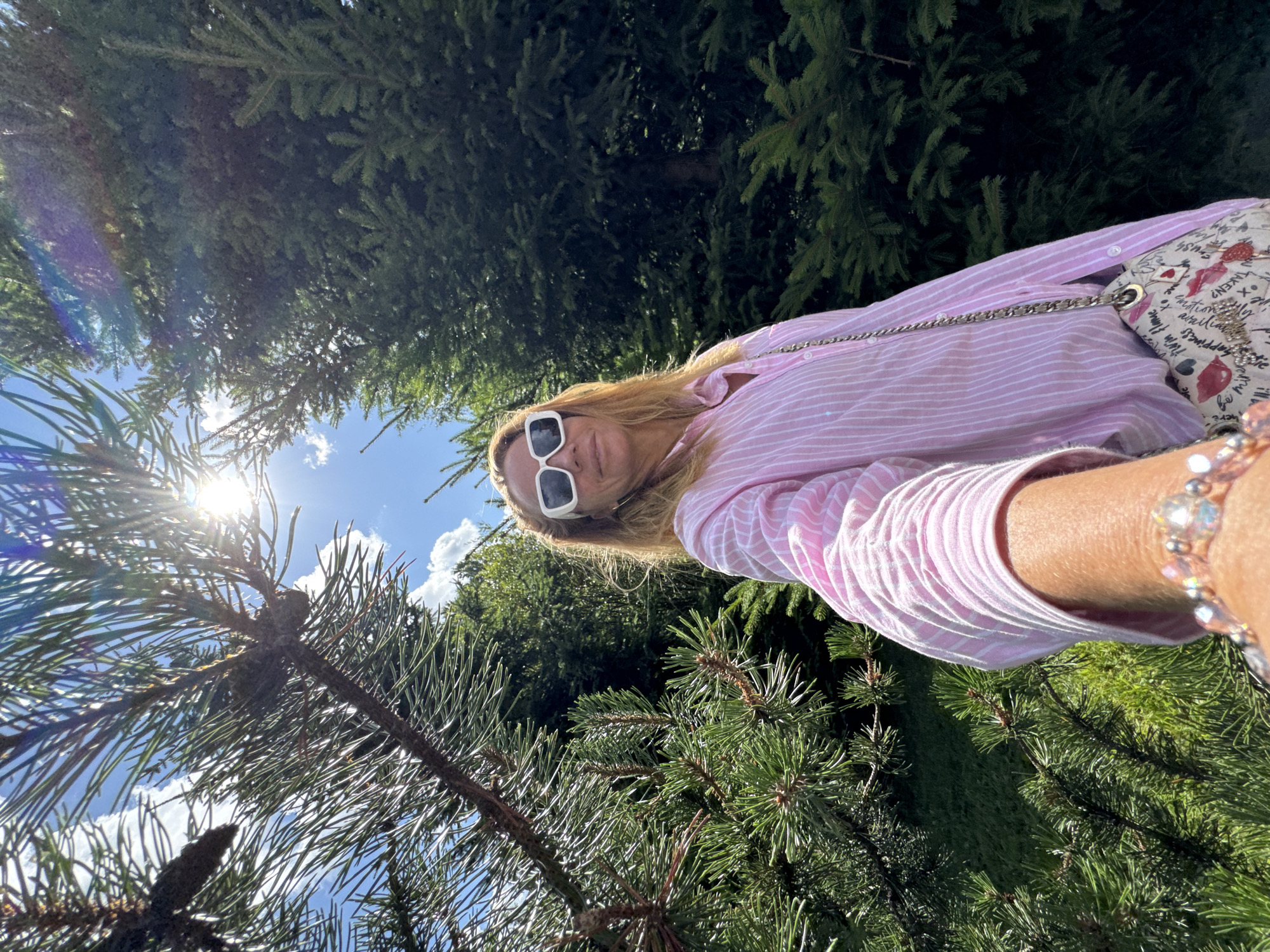 The image shows a woman taking a selfie outdoors, framed by pine trees. The sun is bright and positioned high in the sky, partially obscured by branches. The woman is wearing a pink and white striped button-down shirt, white sunglasses, and a bracelet. She is carrying a light-colored purse with red lips and handwritten text on it. The background consists of lush green pine trees and a clear blue sky with scattered white clouds. The overall scene suggests a sunny day in a natural, possibly park-like setting, given the prevalence of evergreen trees. The location is Sinyavino, Russia.