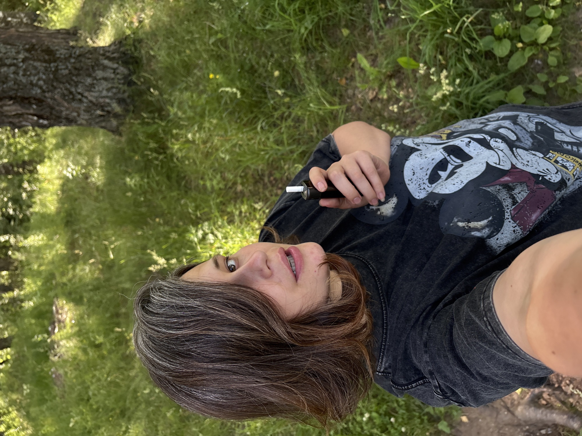 A person is taking a selfie outdoors in a grassy area with trees. The person has brown hair with streaks of grey and is wearing a dark t-shirt with a graphic print of Mickey Mouse. They are holding a dark-colored electronic cigarette device in their right hand. The background consists of green grass, trees, and dappled sunlight. The image was taken in Chaikovsky, Russia.
