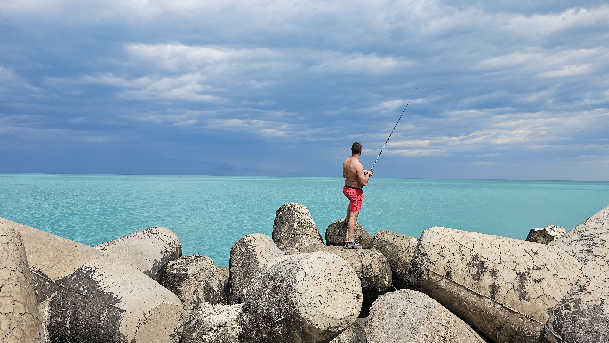 A wide, eye-level shot depicts a man fishing on large, weathered concrete blocks in Varna, Bulgaria. The man, shirtless and wearing red shorts and grey shoes, is viewed from the rear and stands facing the bright turquoise sea, holding a fishing rod. The sky above is cloudy with darker clouds threatening in the distance. The concrete blocks are cracked and stained, creating a textured foreground. The composition emphasizes the contrast between the man's figure, the sea, and the sky.