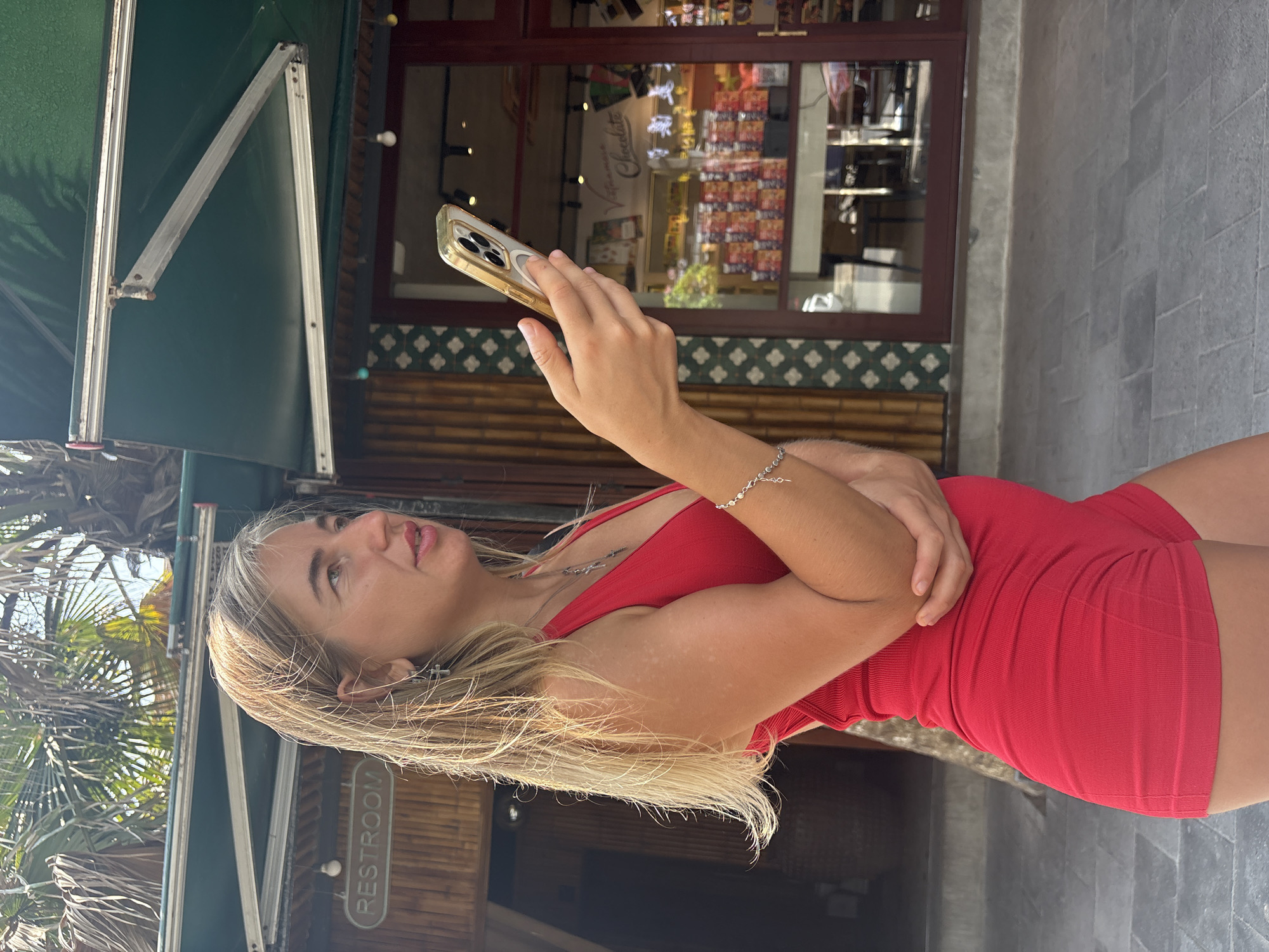 A woman with blonde hair and a red outfit is holding a smartphone in front of a shop in Đà Nẵng, Vietnam. The storefront features a dark wooden frame and glass windows displaying various products. A green awning is visible above the shop. The woman is wearing a bracelet and necklace. The background includes tropical vegetation. The word "RESTROOM" is displayed on a nearby sign. The sidewalk appears to be made of gray stone.