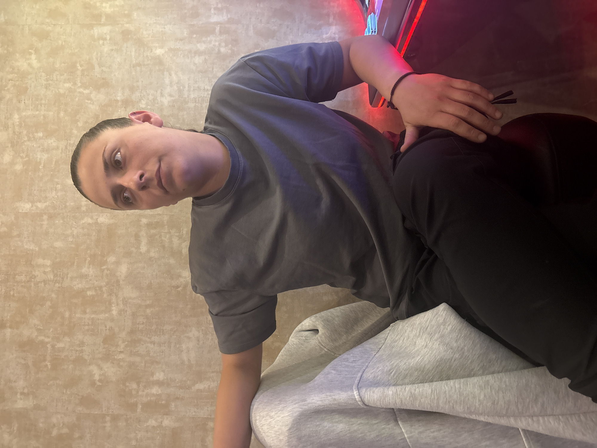 A person is seated indoors, leaning to the right. They are wearing a grey t-shirt and black pants, and a grey hooded sweatshirt. The person has dark hair pulled back and is looking at the camera. A thin black bracelet is visible on their left wrist. The background includes a textured wall and a red lit object, possibly a gaming machine. The location is unknown.