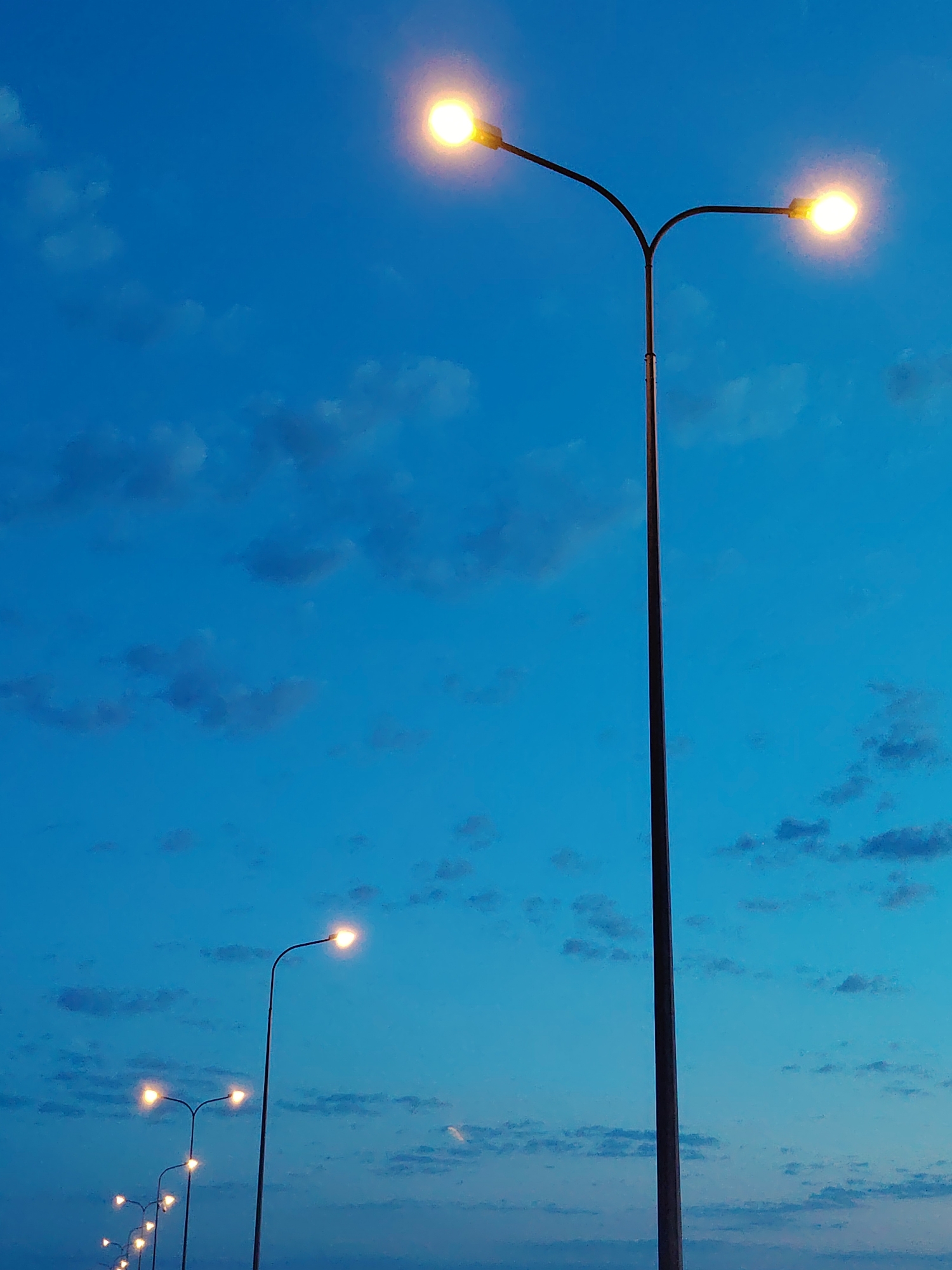 The image shows a row of streetlights against a blue sky at dusk in Chaikovsky, Russia. The foreground features a tall streetlight with two illuminated lamps, while smaller streetlights recede into the distance. The sky is a gradient of blue with scattered clouds.