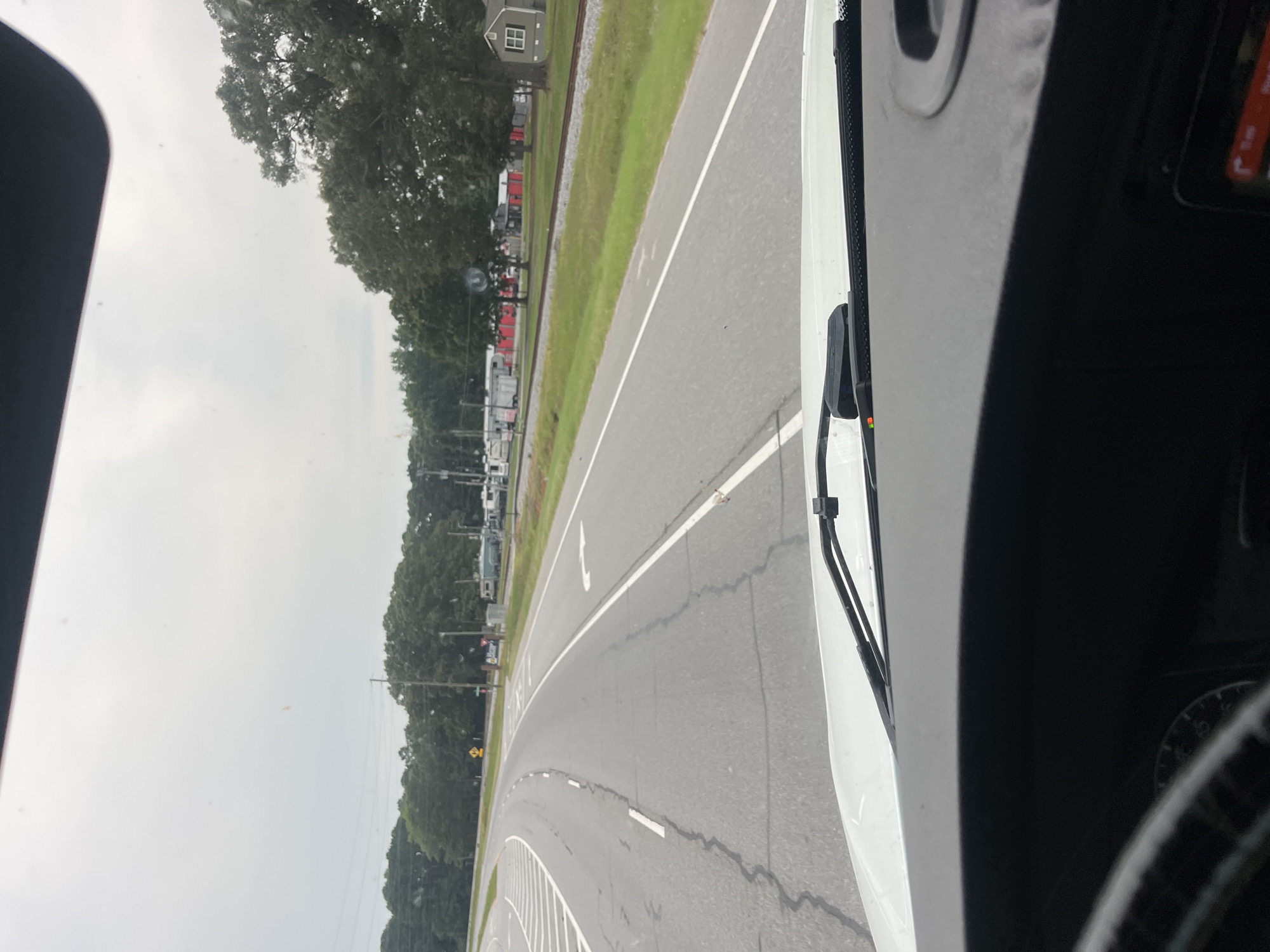 The image shows a view from inside a vehicle looking out through the windshield. The vehicle is on a road with multiple lanes, indicated by white lane markings. The road curves to the left in the foreground. On the right side of the road, there is a grassy area with train tracks running parallel to it. Beyond the tracks, there are several buildings, some of which appear to be storage units or small businesses with red siding. Further in the distance, there are trees and a cloudy sky. The vehicle's dashboard is partially visible at the bottom of the image, with a steering wheel on the left. There are no people visible in the image. The scene suggests a rural or semi-rural area.