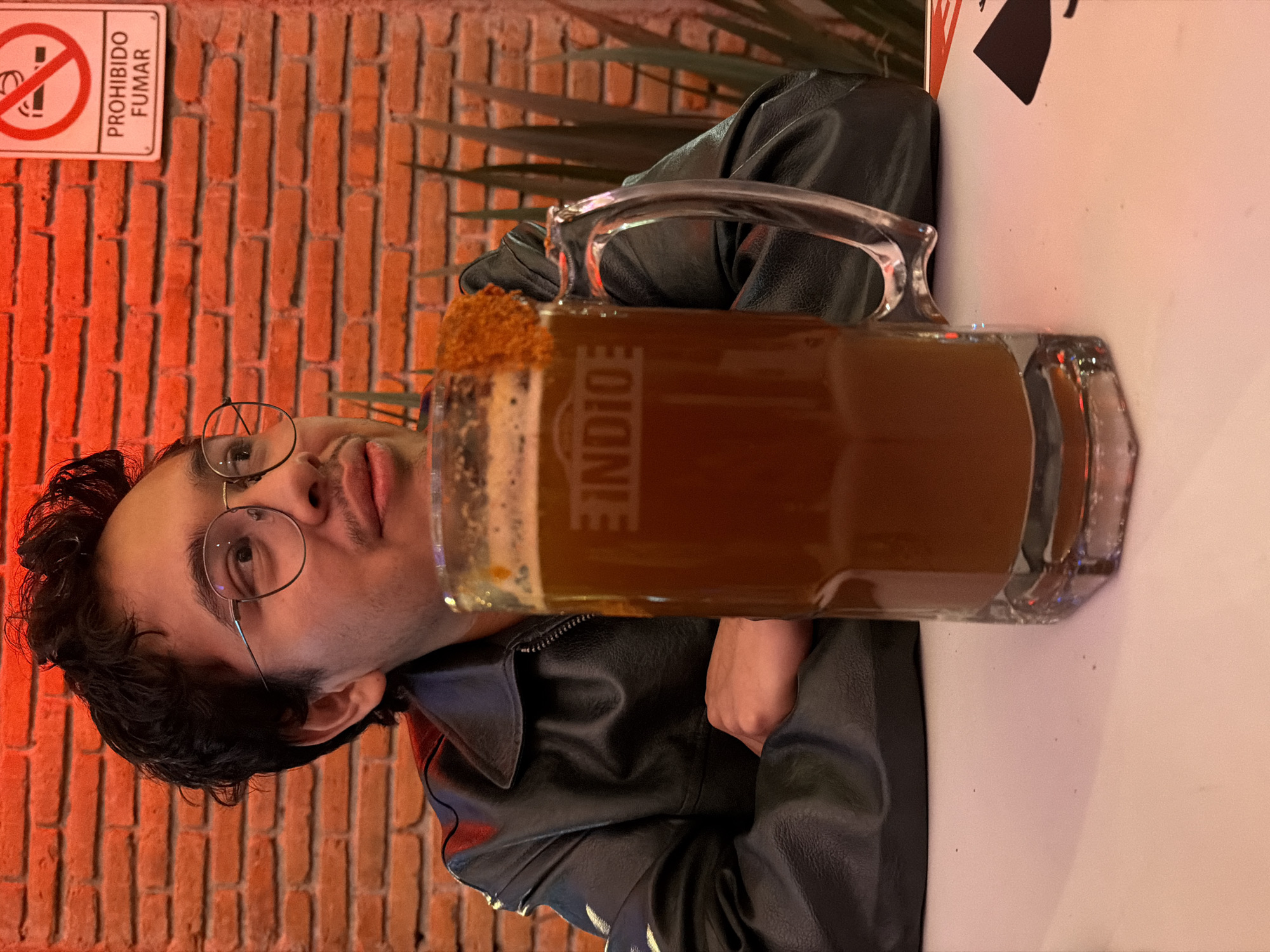 A young man wearing glasses and a black leather jacket is seated at a table with a large mug of Indio brand beer. The beer mug has a chili-salt rim. The background shows a red brick wall and a "Prohibido Fumar" (No Smoking) sign. There are also some plants visible. The location is likely San Andrés Cholula, Mexico.