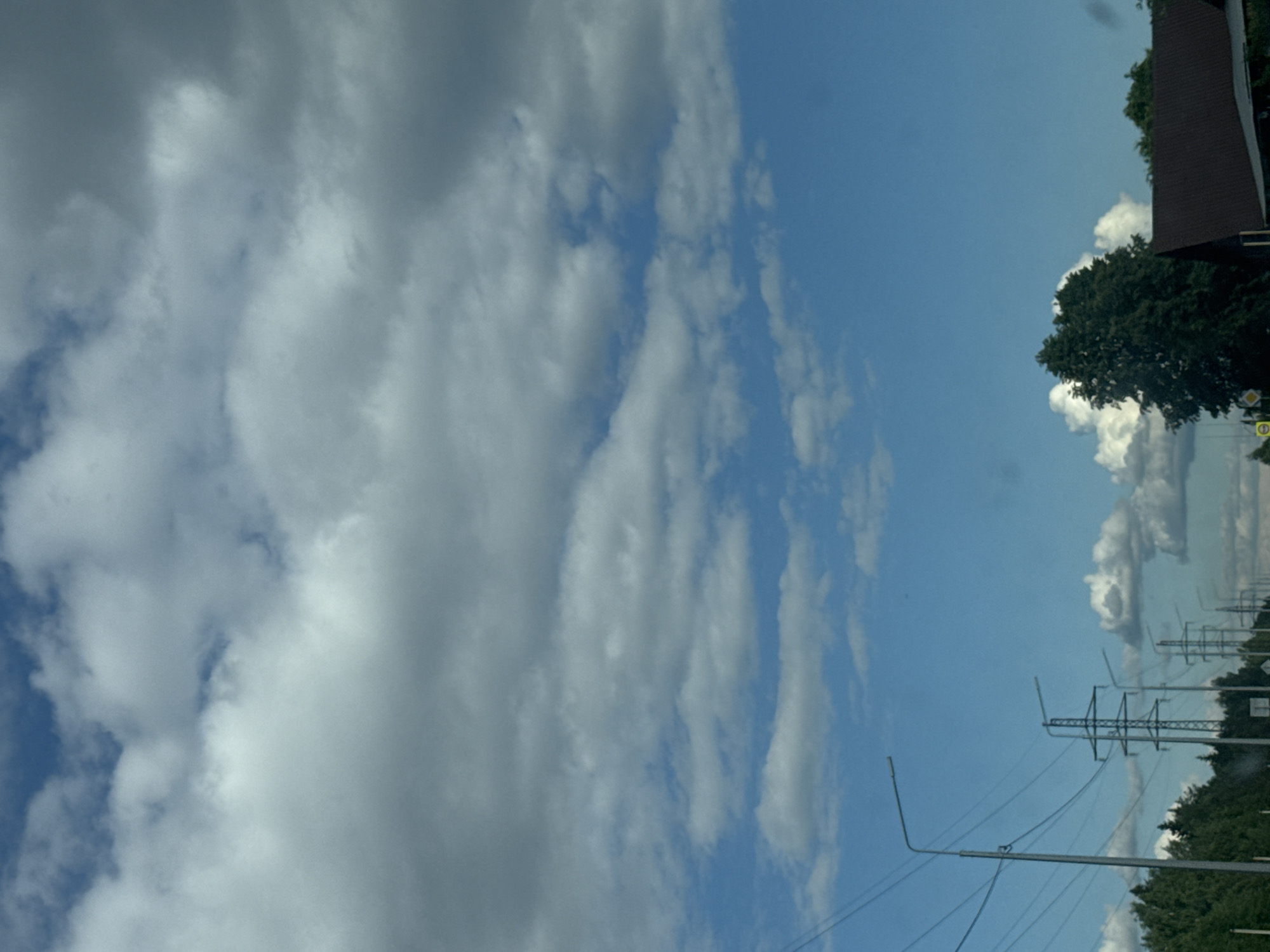 The image shows a cloudy blue sky over a rural or suburban landscape. In the foreground, a line of tall electricity pylons stretches into the distance, with power lines running between them. To the left, a street lamp is visible. A dense line of green trees runs along the horizon, and on the right side of the image, a dark brown roof of a building is visible. There are no people or animals in the frame, and no visible signs of immediate activity. The scene appears to be a road passing through a semi-developed area on a cloudy day. This view is consistent with a location in or around Chaikovsky, Russia, given the presence of infrastructure typical of the region and the overall landscape.