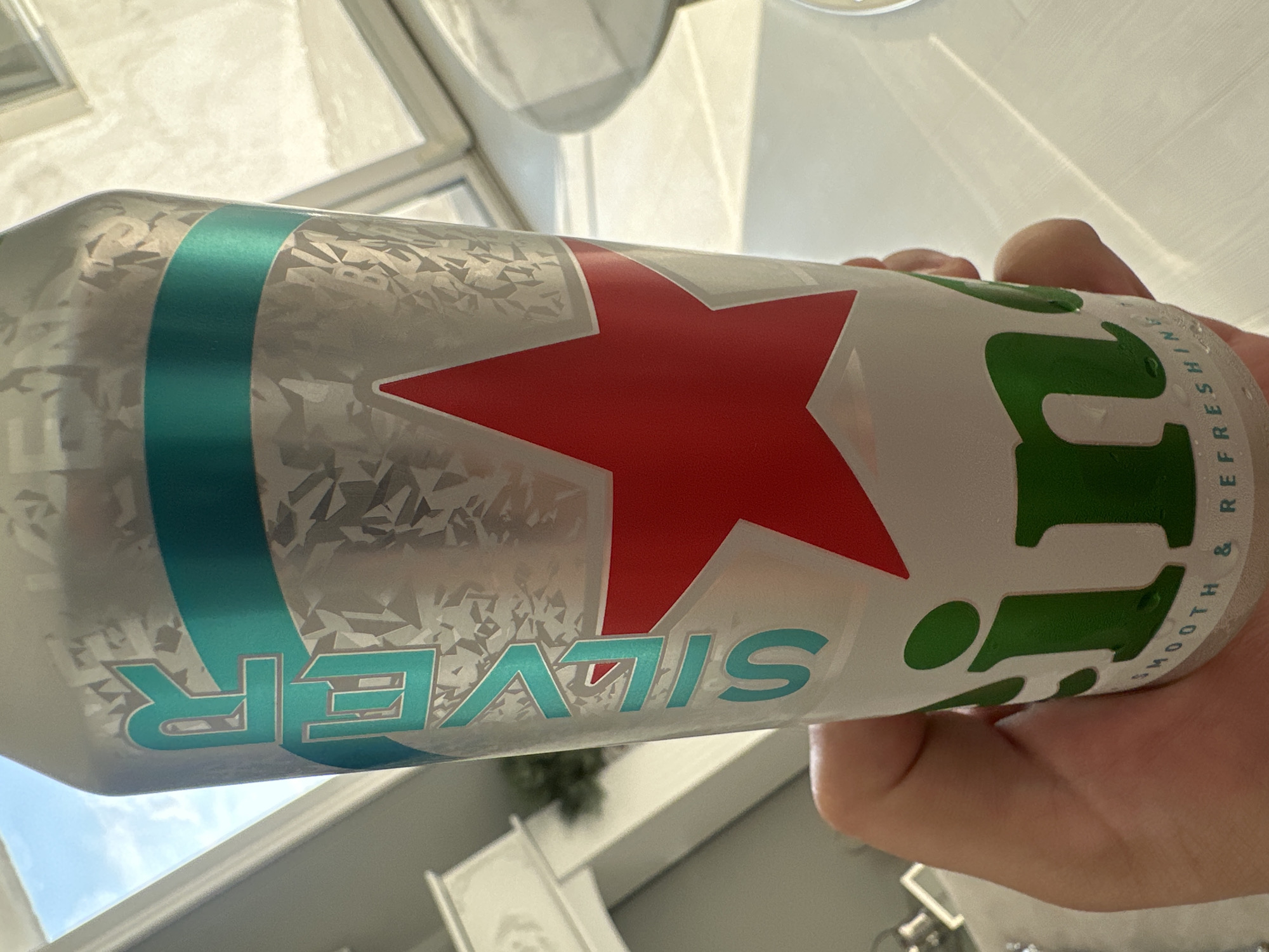 The scene is located in the city of Asenovgrad, Bulgaria. A human hand, with visible skin folds and texture, grasps a tall, slender aluminum beverage can. The can is primarily silver with a textured, faceted pattern resembling ice crystals. Teal-colored capital letters spelling "SILVER" are vertically oriented on the left side of the can, outlined in silver. A solid horizontal teal band crosses the upper middle section of the can. Below this, a prominent red five-pointed star is displayed on a white background. The lower portion of the can reveals two green letters, "i" and "n", part of the "Heineken" brand name, with the phrase "SMOOTH & REFRESHING" printed in small white capital letters along the bottom rim. Condensation droplets are evident on the can's surface. The background depicts a brightly lit indoor environment. A window in the upper left corner frames a clear blue sky with sparse white clouds. White window frames and other white architectural elements are discernible. Sunlight casts distinct linear and geometric shadows across light-colored interior surfaces, likely a table or countertop. A faint green object, possibly foliage, is partially visible in the distant left background.