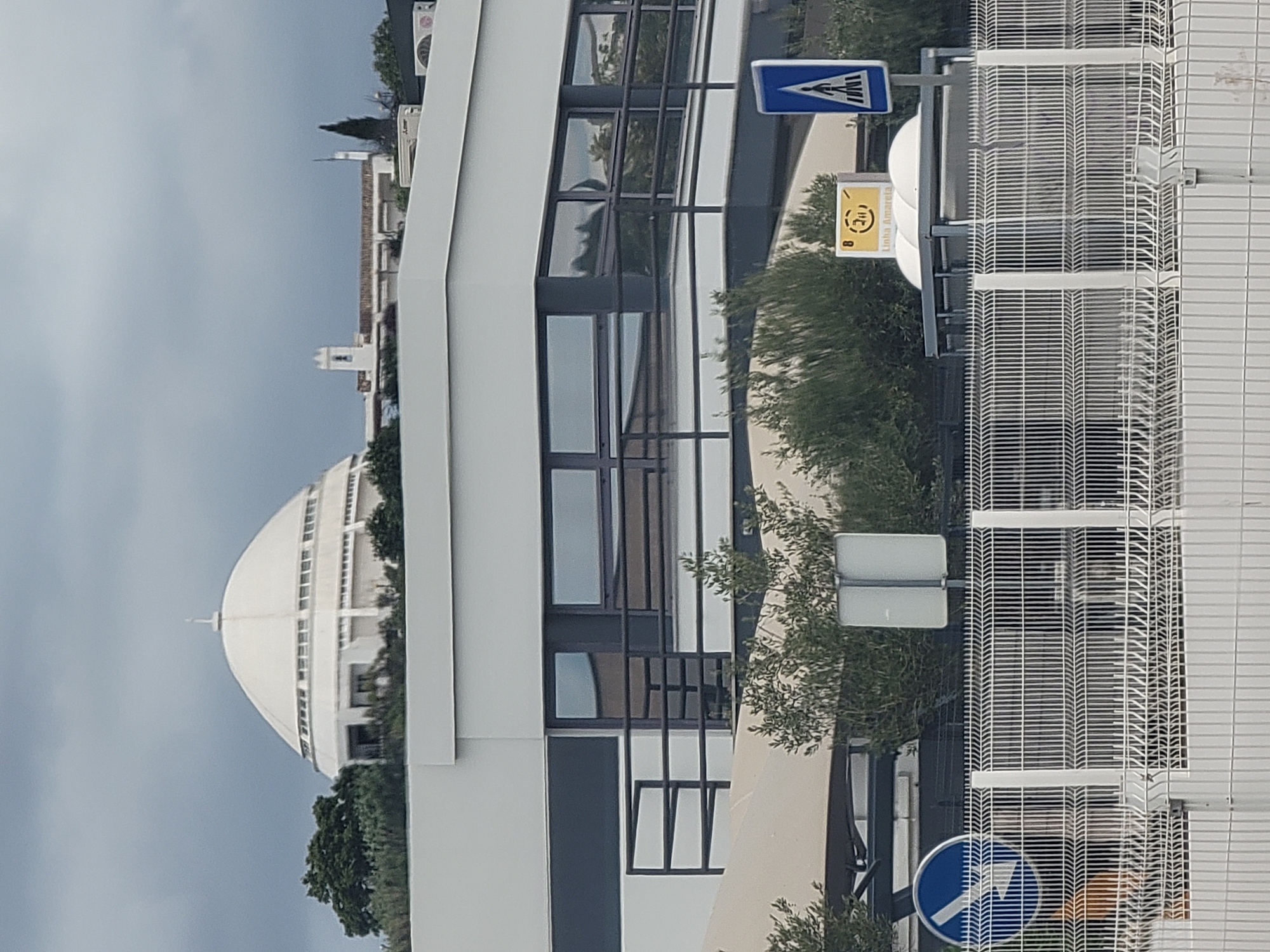 The image shows a modern building with a white, rounded dome structure in the background. The building has large windows and a dark railing. In the foreground, there is a white metal fence. Several road signs are visible, including a blue sign with a pedestrian crossing symbol and a yellow sign with the text "Linha Amarela" (Yellow Line). The scene appears to be in an urban or suburban area, possibly a public space or a commercial district. The sky is overcast. The architecture suggests a contemporary design, possibly a public building or facility. The presence of road signs indicates a functioning road network and pedestrian access.