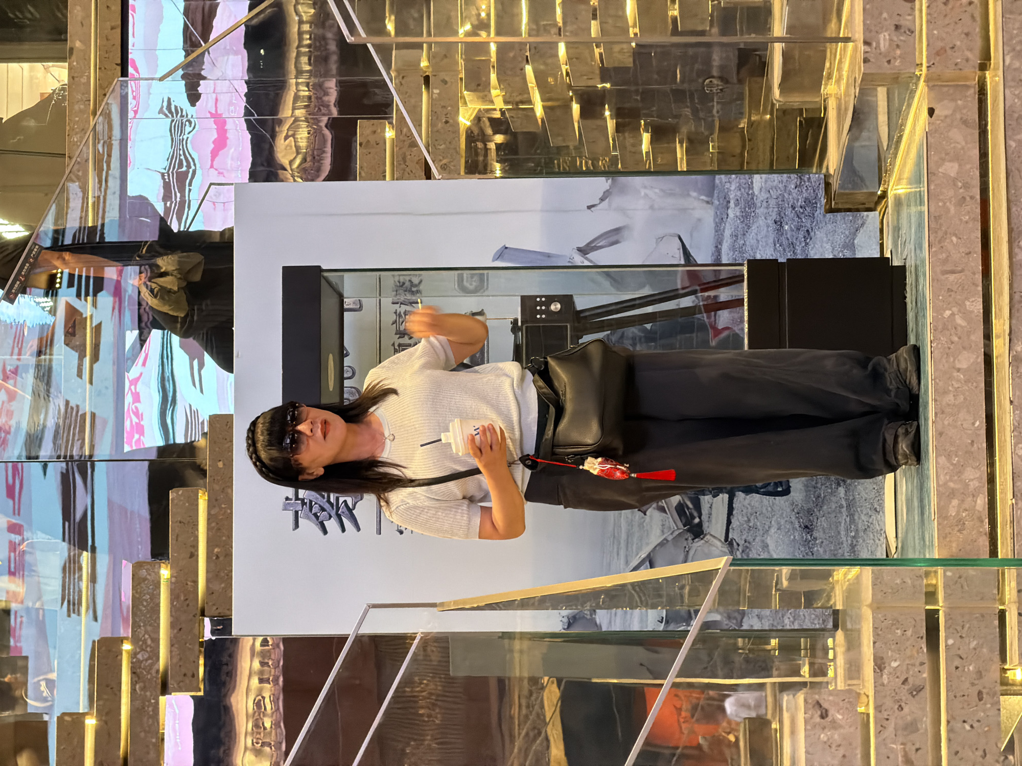 A woman with dark hair, wearing sunglasses and a light-colored short-sleeved shirt, dark pants, and dark shoes, stands on a tiered platform in what appears to be an indoor setting. She is holding a white cup with a black straw in her right hand and has a black purse with a red tassel hanging from her left hip. The background features a large poster with Chinese characters and an image, and to the right, a staircase made of reflective golden blocks and glass railings. The environment suggests a commercial or public space, possibly in Yuzhong District, China, given the context of the poster. The overall lighting is warm, highlighting the reflective surfaces of the staircase and the poster.