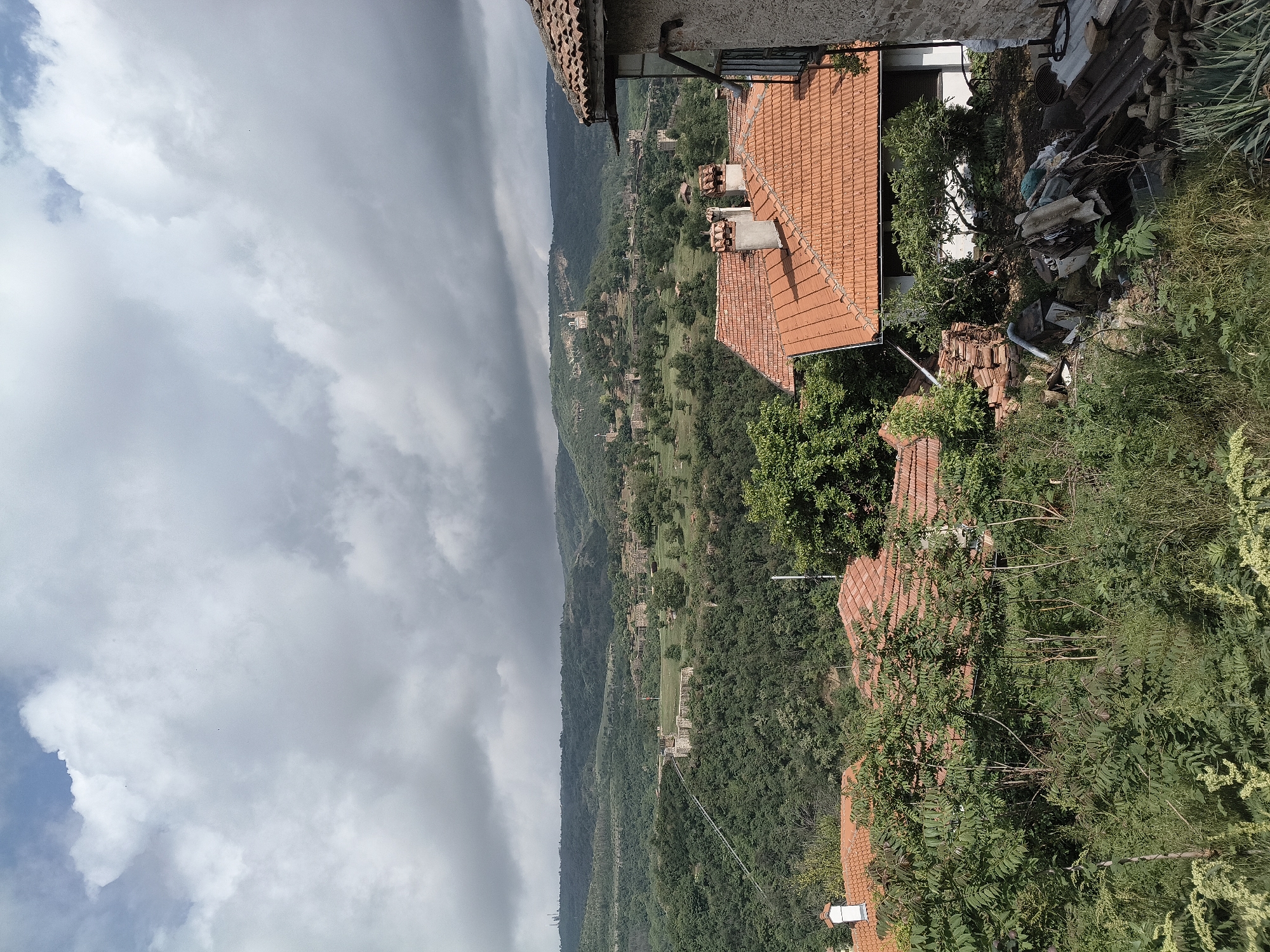A landscape view is presented from an elevated position, capturing a hillside scene with rooftops in the foreground and a mountain range receding into the distance beneath a cloudy sky.
Foreground: A section of a building with a traditional terracotta-tiled roof and possibly a chimney is visible. Adjacent to the building, there is dense vegetation, including trees and shrubs. Scattered debris or construction materials are present, including bricks, metal sheets, and other miscellaneous items.
Midground: The hillside is covered in a thick forest. Interspersed within the trees are clearings or fields. There are some visible structures or buildings, suggesting a settlement or village.
Background: A mountain range extends into the distance. The sky is overcast with a mix of light and dark clouds, hinting at an approaching storm.
The image suggests a rural setting, possibly within the vicinity of Veliko Tarnovo, Bulgaria, known for its picturesque landscapes and historical architecture. The specific location cannot be pinpointed without additional landmarks or contextual clues. No people, actions, or interactions are observed in the image.
