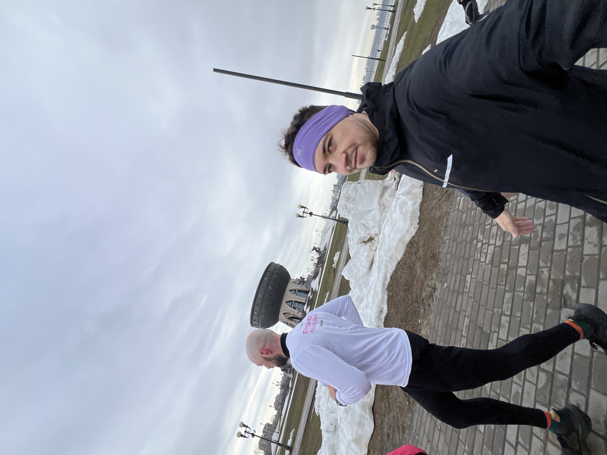Two men stand on a brick-paved path in Kazan, Russia. One man wears a black jacket, a purple headband, and stands in the foreground, facing the camera. Behind him and to the left, the second man wears a white long-sleeved shirt, black leggings, and faces away from the camera. Patches of snow are scattered on the grass alongside the path. In the background, buildings and street lamps are visible. The sky is overcast. The Kazan Family Center is in the background.