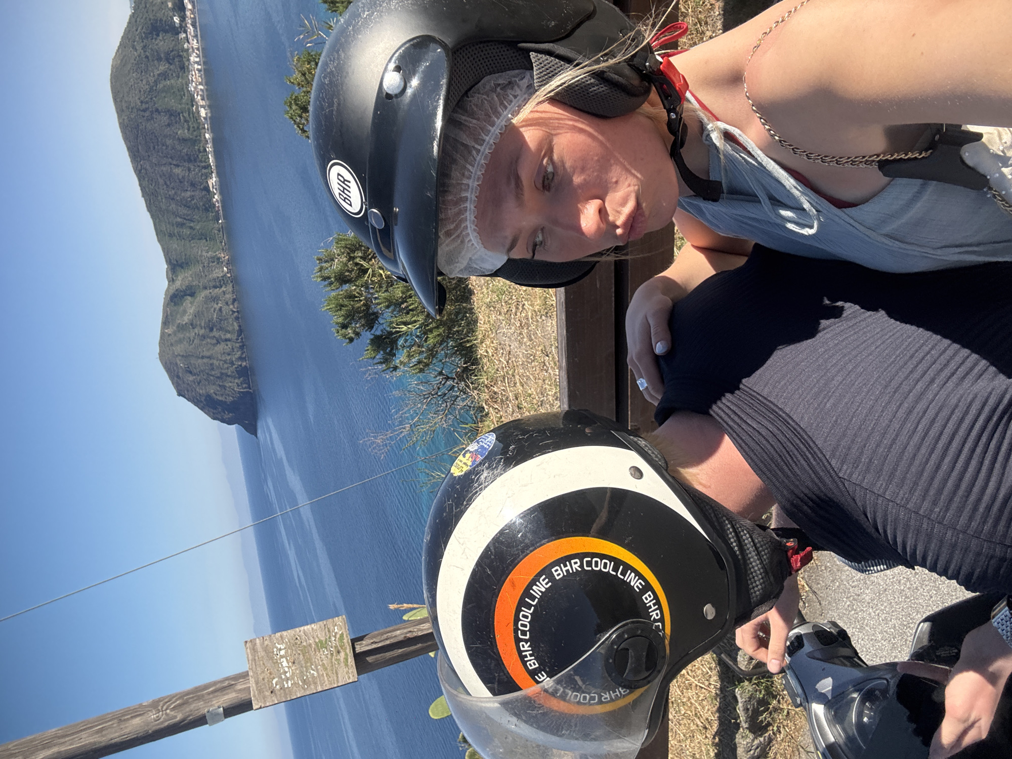 This image captures two individuals, likely tourists, on a scooter or motorbike stopped at a scenic viewpoint overlooking the sea and a mountainous coastline. The photo is tilted, presenting the horizon vertically, but depicts a bright, sunny day with a clear blue sky. The setting, identified as Lipari, Italy, features deep blue waters and verdant, rugged hillsides with some scattered buildings visible along the distant shore.
The person in the foreground, a blonde woman, is seen wearing a black open-face helmet with a "BHR" sticker, over a white disposable hair net. She has a slight pout on her face and is dressed in a light-colored, possibly light blue, strappy top, with a red bra or bikini top visible underneath. A golden-colored chain strap runs over her shoulder. Her hand, with light nail polish, rests on the arm of the person next to her.
The second person, presumably the driver, is partially visible at the bottom of the frame, also wearing a black open-face helmet. This helmet features a distinctive circular design with orange and white stripes and the repeated text "BHR COOLLINE." This individual is wearing a dark, ribbed top and their hands are on the handlebars of the scooter, which is partially visible.
In the mid-ground, a wooden fence or railing separates the subjects from the vast expanse of the sea. To the left, a weathered wooden sign on a post is visible, though its text is too faded to be legible. The overall impression is a leisurely stop on a coastal road during a sunny, warm day, enjoying the panoramic view.