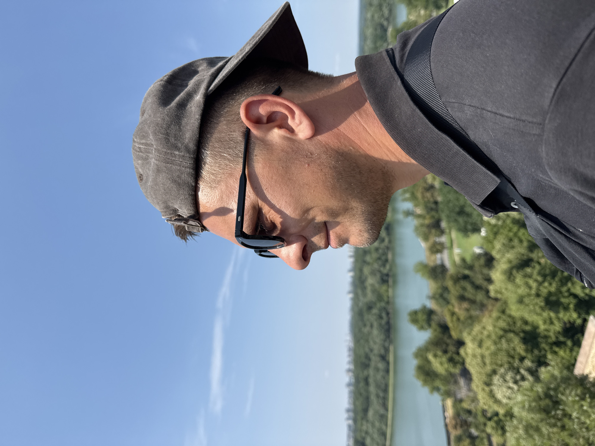 The image shows a close-up, side profile of a Caucasian man wearing a dark gray baseball cap and dark sunglasses. He has short, light brown hair and is looking towards the left of the frame. He is wearing a dark gray polo shirt.
The background features a wide, calm river, with a city skyline visible in the distance across the river. Lush green trees and vegetation are abundant in the foreground and middle ground. The sky is clear and bright blue with a few wispy clouds. This scene is consistent with a viewpoint from Kalemegdan Fortress in Belgrade, Serbia, overlooking the confluence of the Sava and Danube rivers and the New Belgrade cityscape.