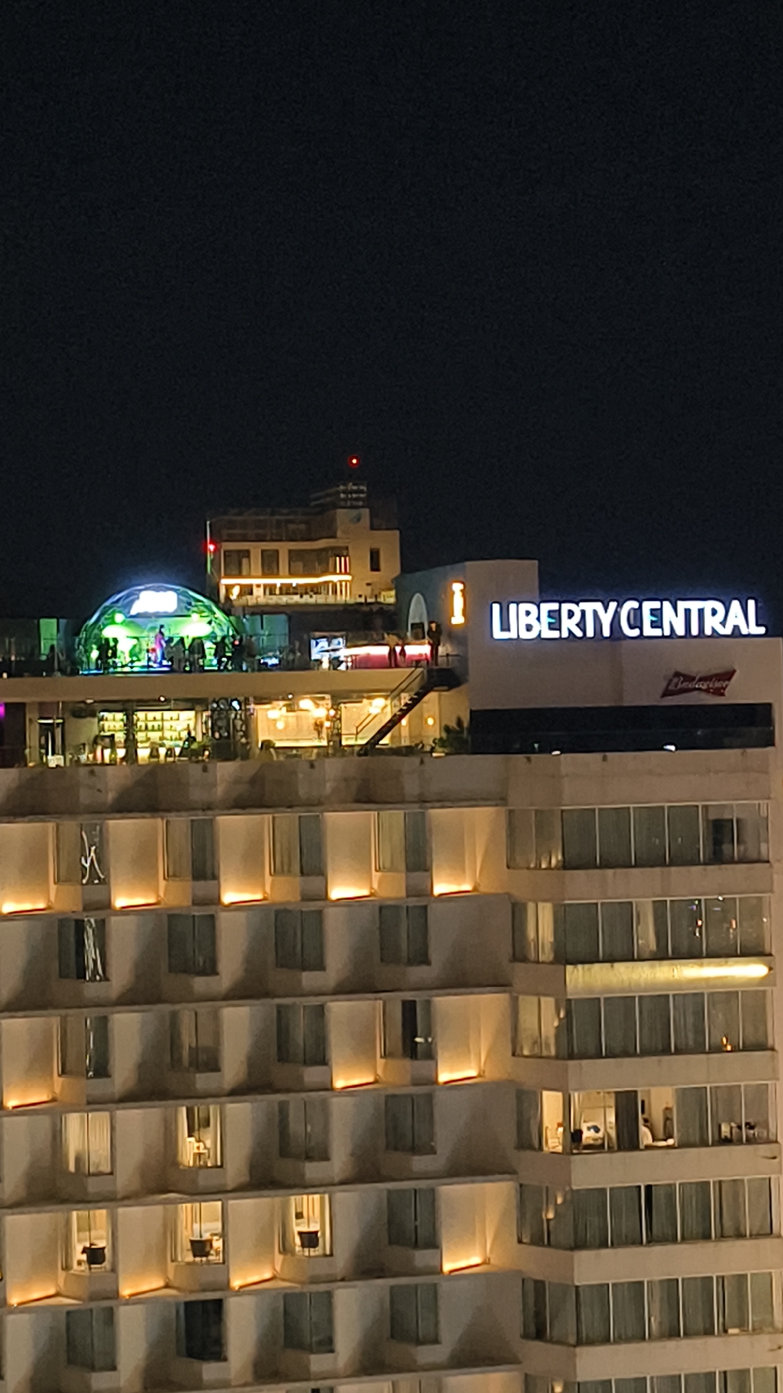 The image captures a nighttime view of the Liberty Central Nha Trang Hotel in Nha Trang, Vietnam. The hotel building dominates the frame, with its facade illuminated by external lights, highlighting rows of windows. On the upper levels, a rooftop area is visible, featuring a large, glowing geodesic dome, likely housing a bar or event space, with silhouetted figures visible inside. Adjacent to the dome, a bar setup with illuminated shelves and seating can be seen. A staircase leads up to this level. A prominent sign on the building reads "LIBERTY CENTRAL" in large, illuminated letters. A smaller Budweiser logo is also visible. The dark night sky serves as the background.