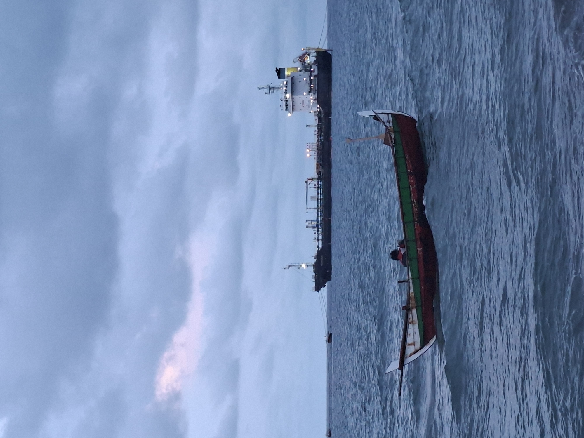 The image shows a fishing boat with two people on board in the foreground. A large oil tanker is docked in the background, connected to land by a series of pipes and walkways supported by pylons. The sky is overcast. The location is most likely in the city of Lhokseumawe, Indonesia.