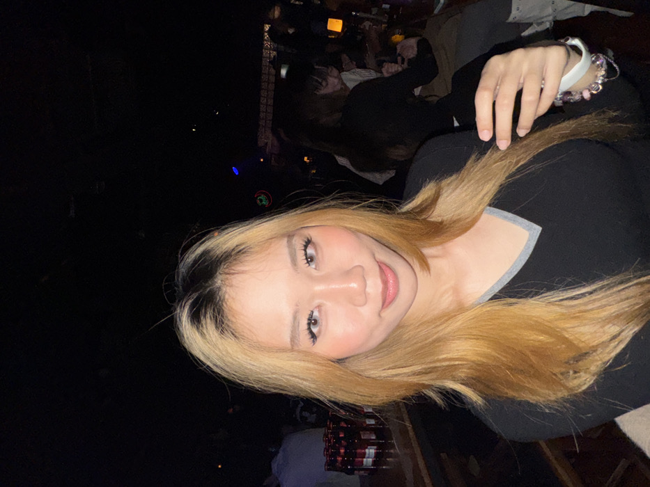 The image displays a young adult female positioned centrally in a dimly lit indoor environment, consistent with a bar or nightclub. She has long, layered hair with dark roots transitioning to blonde, and wears a black V-neck top with a grey trim. Her eyes, enhanced with light-colored contact lenses and visible eyelashes, are directed towards the camera, displaying a slight smile. A white smartwatch and a charm bracelet with various pink and purple elements are present on her left wrist. Her right hand is positioned near her chest, lightly touching her hair.
The background features a wooden bar counter on the left, displaying multiple bottles with red labels arranged in rows. A partially visible individual in a white shirt stands behind these bottles. The right and distant background reveals an array of indistinct patrons seated at tables, largely obscured by the low light. One female figure in a dark top is more discernible in the mid-ground, seated at a table. Ambient lighting sources, including a faint blue light and a red circular light, are visible overhead. The specific location within the city of Liangjiang, China, is not discernible from the visual information provided.