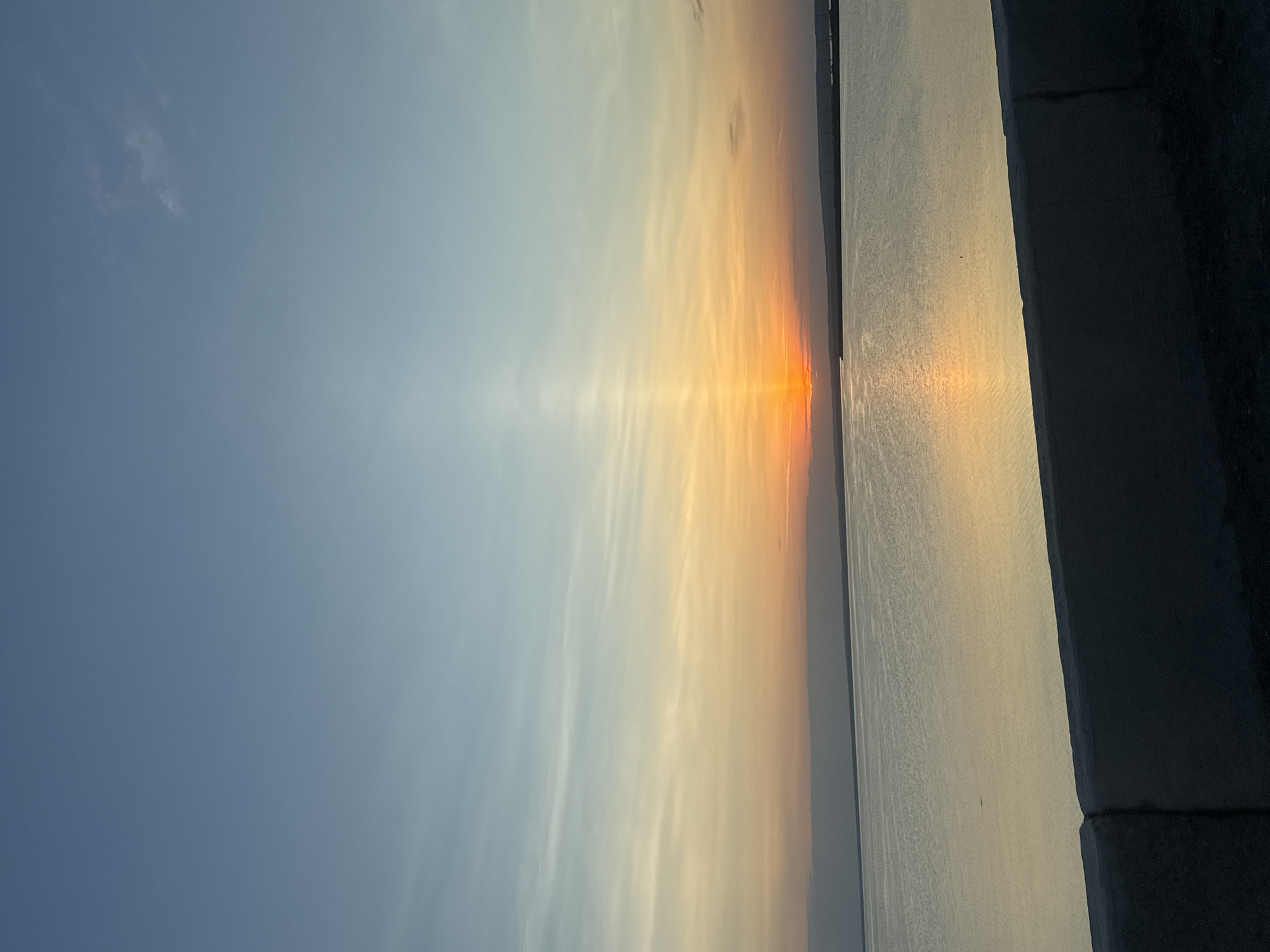 The image depicts a sunset over water, likely the Kama River, near Chaikovsky, Russia. The sun is positioned low on the horizon, casting a bright orange glow across the water and the sky. The sky transitions from a deep blue at the top to a light orange near the horizon, with some wispy clouds present. The water reflects the sunlight, creating a shimmering effect. A concrete structure is visible in the foreground. There are no people or other distinct objects visible.
