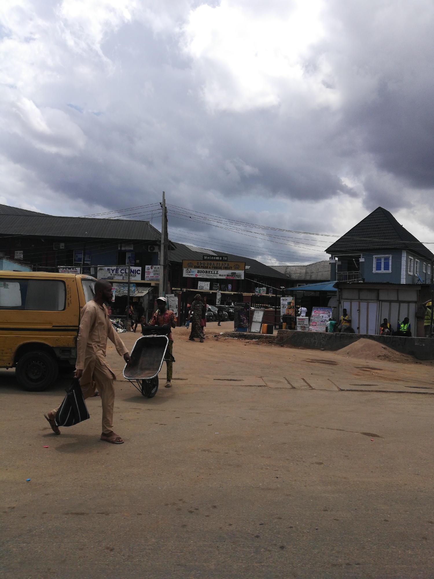 The image captures a scene in Egan, Nigeria, characterized by a street intersection bustling with activity under a cloudy sky. A yellow van is parked on the left side of the image, with a man in traditional attire walking past, carrying a bag in one hand and pushing an empty wheelbarrow with the other. Another individual assists with the wheelbarrow. The foreground is dominated by a paved road with visible wear.
In the background, there is a multi-story building with commercial establishments like an "EYE CLINIC" and "BIOBELLZ." Signage indicates the presence of "OMO OBA PLAZA" and "CAR PARK & PUBLIC TOILET." There are various people in the background, including some sitting on a raised concrete area.
A two-story blue and white building is on the right side of the image, with some people sitting in front of it. The overall impression is a vibrant, if somewhat chaotic, urban scene.