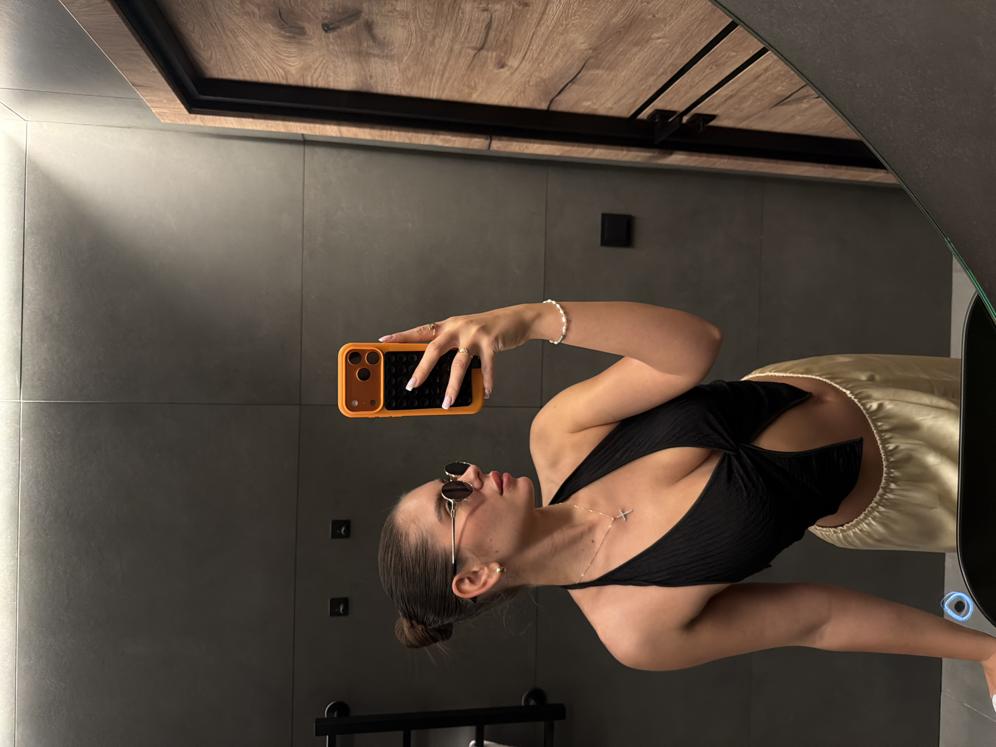 The scene is located in Манский, Russia. A young adult female, positioned for a mirror selfie, is captured from the chest up, slightly angled to her right, looking into her smartphone. She wears dark, round sunglasses with gold frames, a black textured halter-neck top with a deep V-neck and a front twist detail, revealing her midriff, and light golden, loose-fitting pants. Her dark hair is pulled back into a neat bun, and she has a small gold hoop earring on her visible left ear. A thin gold chain with a small silver-colored cross pendant hangs around her neck. On her left wrist, she wears a white beaded bracelet, and on her right hand, two thin gold rings are visible on her index and ring fingers. Her fingernails feature a French manicure. She holds an orange smartphone with a black grid-patterned case in her right hand, its screen dark. The background consists of dark gray, large-format tiled walls with thin grout lines. To the right, a modern wooden door with a dark frame is partially visible. Various dark wall fixtures, including a towel rail on the left and several square elements, are mounted on the tiled surface. The bottom left of the reflection shows a dark countertop and a white sink basin with a glowing blue ring around what appears to be a drain or sensor.