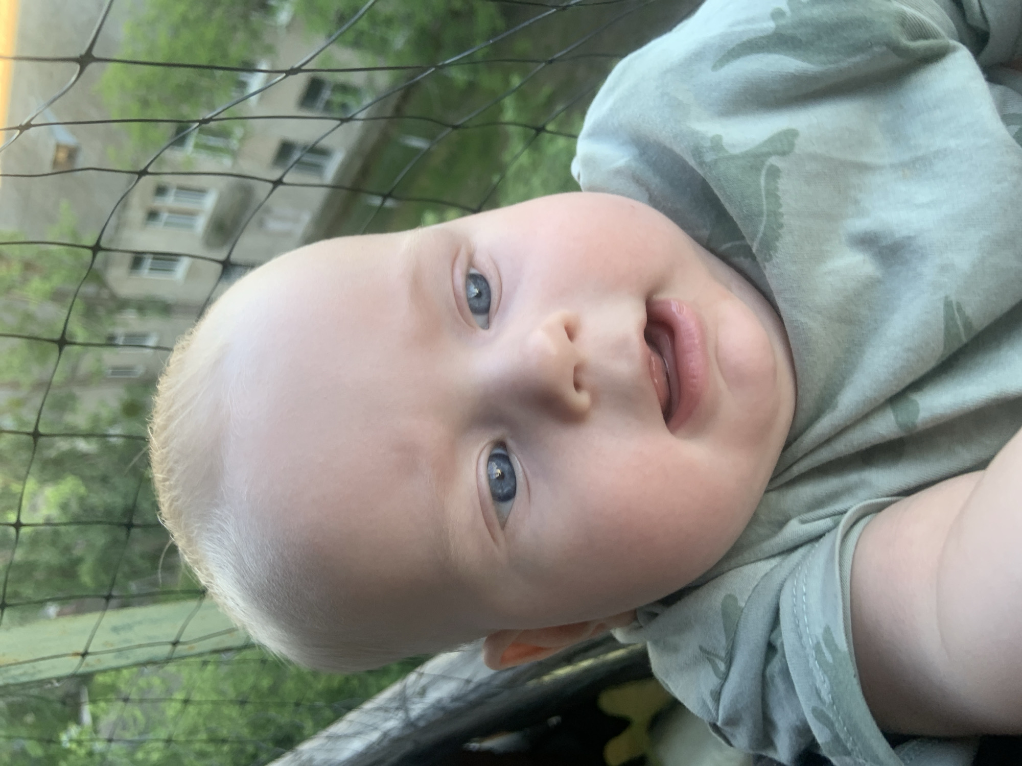 The image shows a close-up, Dutch angle shot of a baby with fair skin and light hair. The baby has blue eyes and is looking upwards. The baby's mouth is slightly open, revealing their tongue. They are wearing a light green t-shirt with a camouflage pattern. A black net is partially visible in the foreground, with a building and greenery in the background, suggesting an outdoor setting. The lighting indicates it might be daytime. There is no visible text in the image. The overall mood is candid and focuses on the baby's features.
