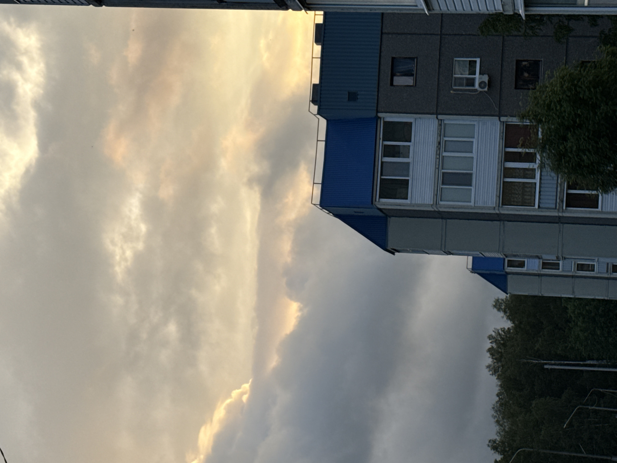 The image captures a section of an apartment building with a blue roof against a cloudy sky. The building appears to be multi-story, with visible windows and balconies. In the foreground, a line of dark green trees is present. Streetlights are also visible on the left side of the image. The sky is filled with clouds, with patches of sunlight breaking through, suggesting either dawn or dusk. The scene is set in a residential area within the city of Марковский, Russia.