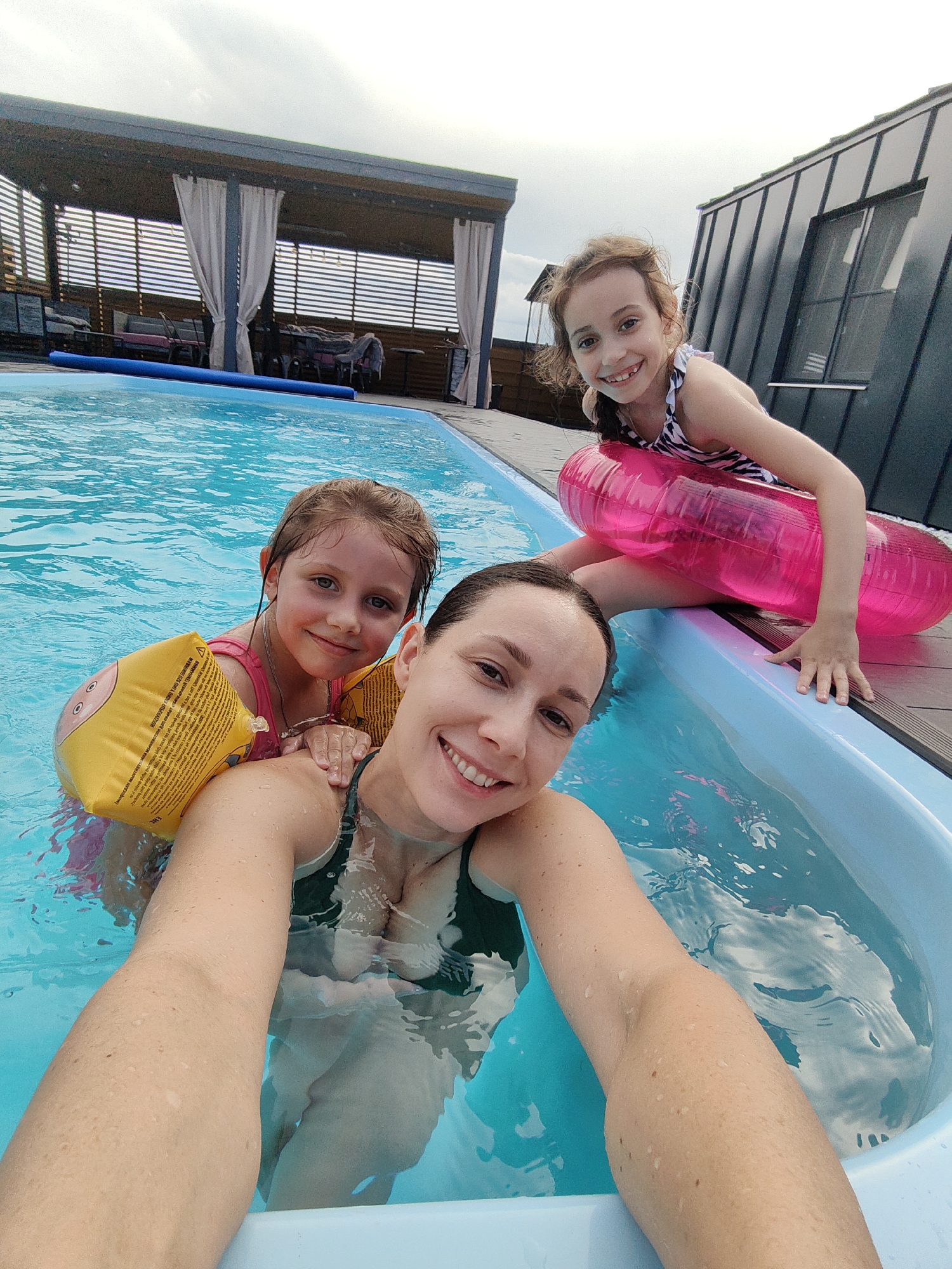 In Zheleznovodsk, Russia, a woman and two young girls are in a swimming pool. The woman is in the foreground, taking a selfie, with one girl partially visible behind her. The second girl is in the background, sitting on the edge of the pool with a pink inflatable ring. The pool area features a wooden deck, a covered patio with furniture, and a modern building with a dark, sloped roof. The sky is overcast.