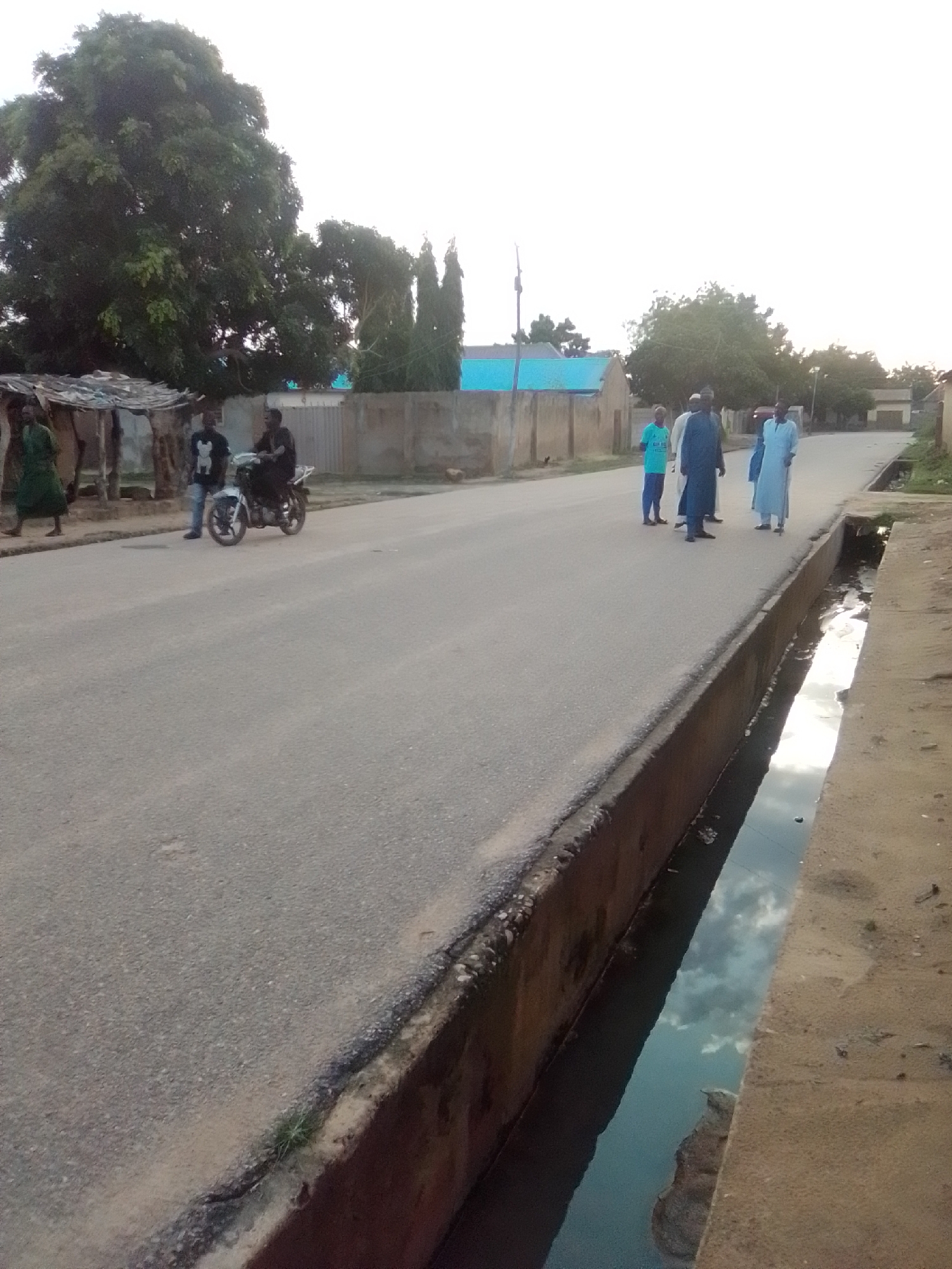 An eye-level, medium shot captures a street scene in Gombe, Nigeria, at dusk. The foreground is dominated by a concrete drainage ditch filled with murky water reflecting the sky. The asphalt road stretches into the background, bordered by a dusty embankment on the right.
On the left side of the road, a person in a green robe walks past a simple, open-sided shelter constructed from wood and corrugated metal. Further down the road, two men are present; one stands next to a white motorcycle while the other sits on it.
Towards the center of the frame, on the road, a group of five men is walking. From left to right, they are dressed in a light blue t-shirt and trousers, a dark blue robe, a light blue robe, and another man in a light blue robe.
The background reveals buildings with concrete walls and blue-tiled roofs, along with a large, leafy tree on the far left. The sky is overcast, indicating the approaching evening. The overall impression is one of a quiet, lived-in urban environment.