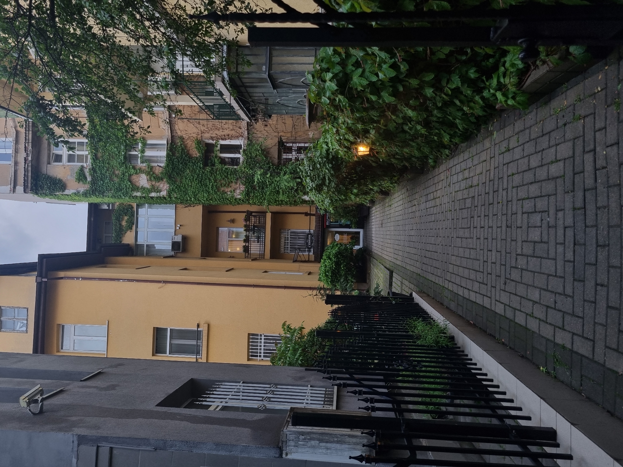 A narrow alleyway, paved with gray rectangular bricks, is lined with buildings and vegetation. A black wrought-iron fence runs along the right side of the alley. The buildings are mostly two to three stories high, painted in various shades of yellow and gray. One building is covered in ivy. The sky is overcast. The location is likely in Sofia, Bulgaria, given the architectural style and vegetation. A lamppost emits a warm glow near the end of the alleyway. There are no people visible in the image.