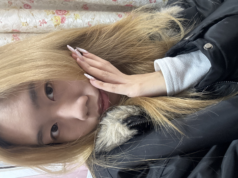 An indoor, close-up portrait captures an individual with fair skin and long, light blonde hair, looking directly forward. The subject wears a black puffer jacket featuring a light brown faux fur trim around the collar or hood. A white, possibly fleece, long-sleeved garment is visible at the left wrist. The subject's left hand is raised, with fingers lightly touching the left side of the face. This hand displays long, pointed artificial nails, white in color with small dark patterns. The background is out of focus, revealing a floral patterned fabric, likely a curtain, with pink and yellow motifs on the right, and a pale pink or white surface on the far left. The specific location within Shapingba District, China, cannot be determined from the visual information provided.