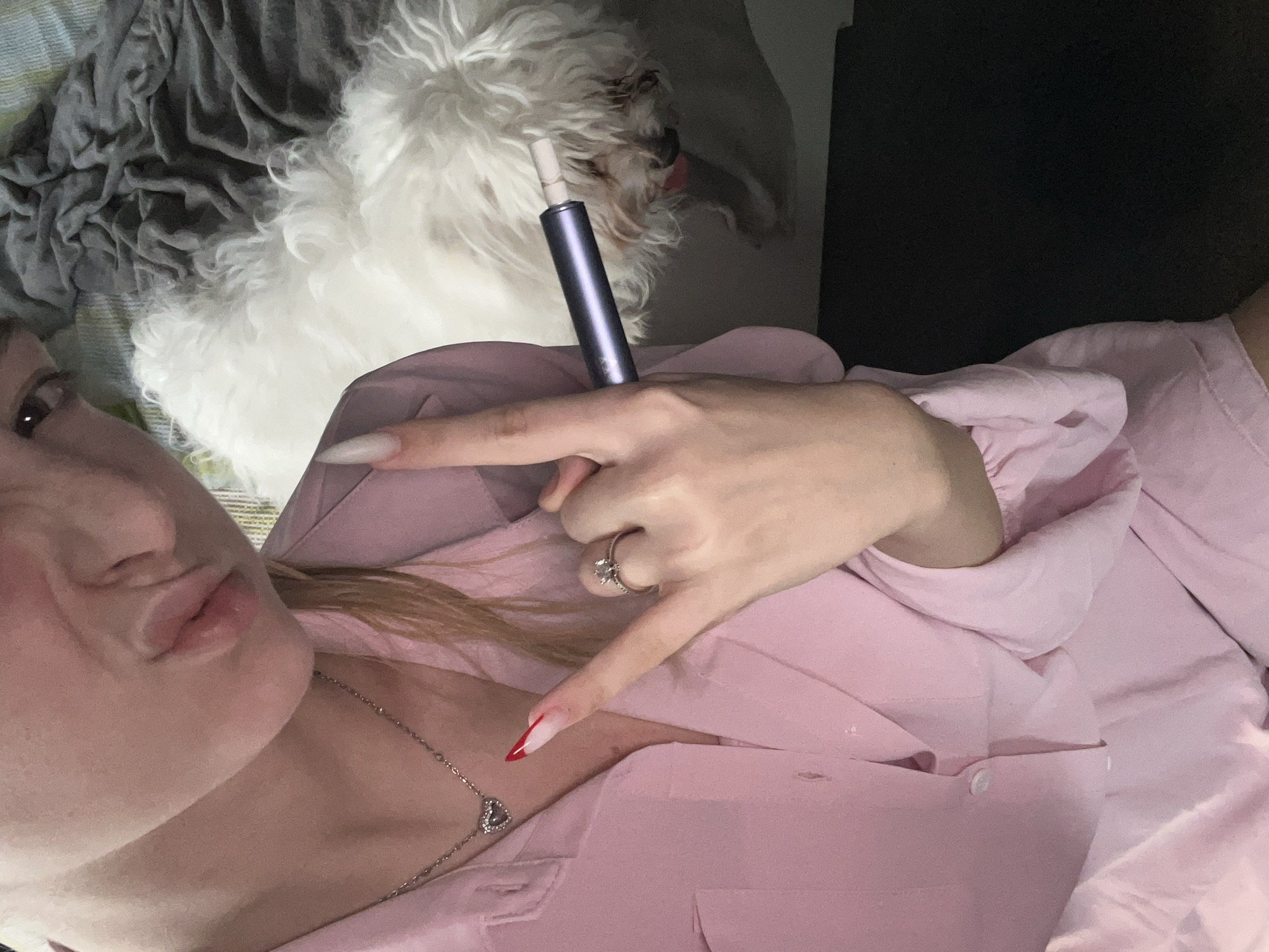 A selfie taken indoors. A woman wearing a pink button-down shirt and a heart necklace, with long fingernails (one has red tips), is posing with a 'rock on' hand gesture. She has fair skin and blonde hair. On the bed behind her is a white fluffy dog and a cylindrical object that appears to be a smoking device. A gray blanket is on the bed, and a dark surface is visible in the background. There is no information about the location in Varna, Bulgaria in the image.