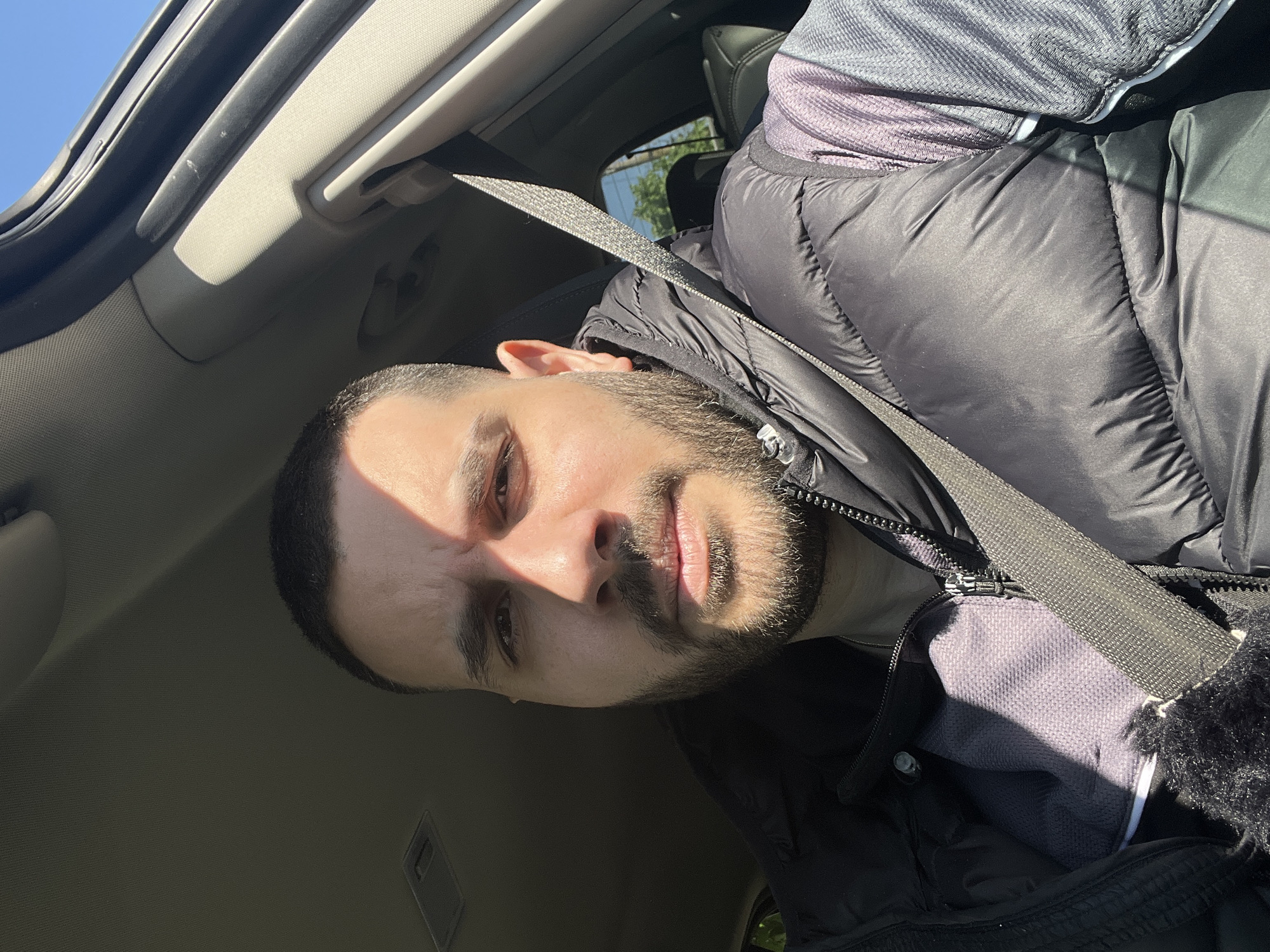 A medium shot depicts a man with short dark hair and a beard, seated in a car. He is wearing a black puffer jacket and a seatbelt. The car's interior is visible, including the ceiling and dashboard. Sunlight is streaming into the car, casting shadows on his face. The location of the photo cannot be verified as Trastikovo, Bulgaria, from this image.
