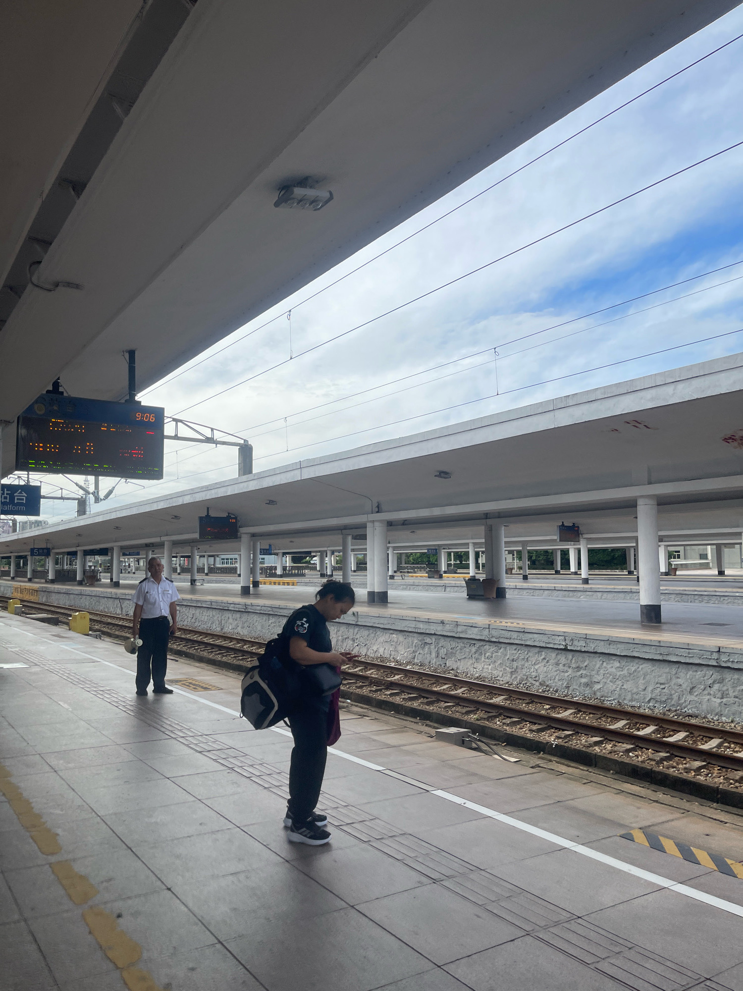 The image captures a train station platform in Baiyun District, China. A man in a white uniform and black pants, holding a round object, stands on the platform. A woman wearing a black shirt and pants, with a backpack slung over her shoulder and a bag in her hand, is also on the platform, looking down at her phone. Two large electronic display boards are visible above the platform, showing train information and the time 9:06. The platform is paved with concrete tiles and bordered by railway tracks. The station features a white roof supported by pillars, with power lines running overhead against a cloudy sky. In the background, another platform with similar architecture is visible.