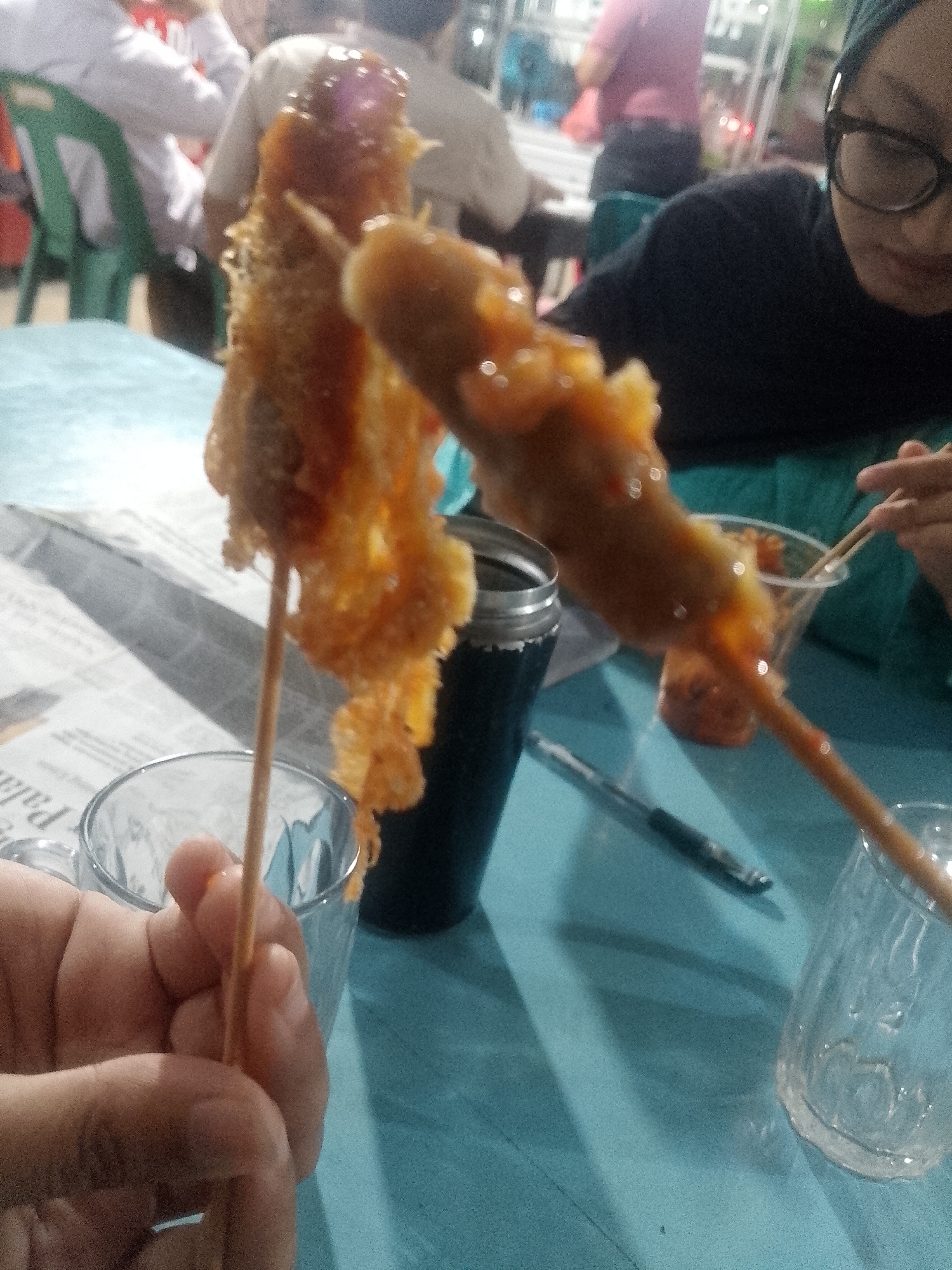 The image shows a close-up of a hand holding a skewer with two fried food items covered in sauce. In the background, blurred figures of people are visible, some seated at tables in what appears to be an outdoor or semi-outdoor food stall setting. A teal-colored table surface dominates the foreground and midground. To the left, a clear glass with a newspaper beneath it is partially visible. To the right, another clear glass and a black thermos or container are also present. A pen lies on the table. The lighting suggests it might be evening or dusk. The overall scene depicts a casual dining experience, likely in Lhokseumawe, Indonesia, given the location context. The focus on the food and the casual atmosphere hint at a street food or market environment. No discernible text is visible in the image.