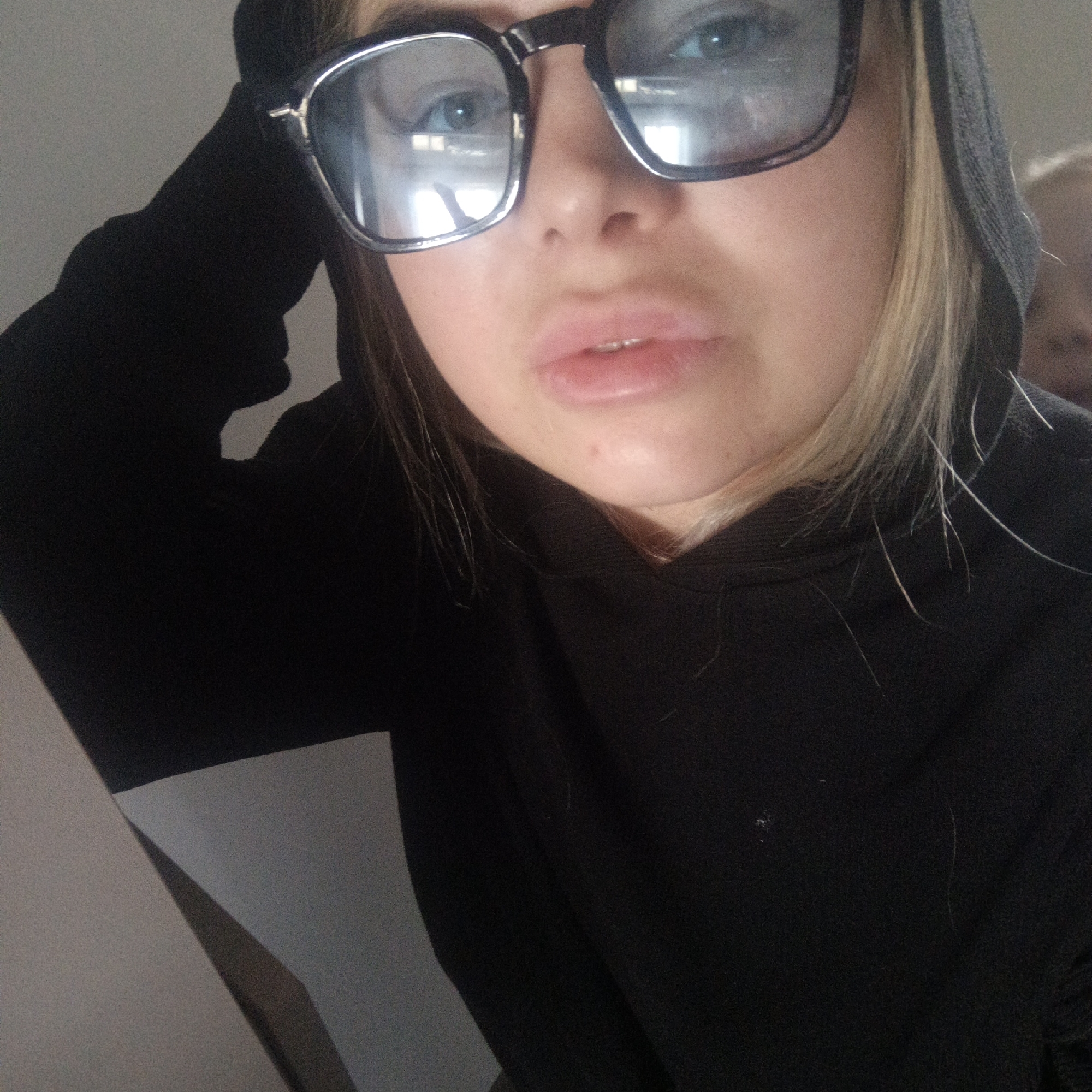 A close-up, chest-up shot features a young person, likely female, looking directly into the camera. They are wearing a black hooded sweatshirt (hoodie) with a light-colored, possibly white or light gray, panel on the left side. Large, rectangular, dark-framed glasses cover their eyes and part of their nose, reflecting light sources from the environment. Through the glasses, blue-green eyes are visible. Blonde hair peeks out from under the hood and frames the face. The person's lips are slightly parted, revealing a hint of their upper teeth. A minor blemish is visible on the chin.
The background is predominantly plain and light-colored on the left, transitioning to a blurry, out-of-focus shape on the right, which suggests another person or object in the distance. The lighting appears to be interior, casting some reflections on the glasses. The overall expression of the subject is neutral. No text is visible in the image. The composition suggests a casual selfie or a close-up portrait.