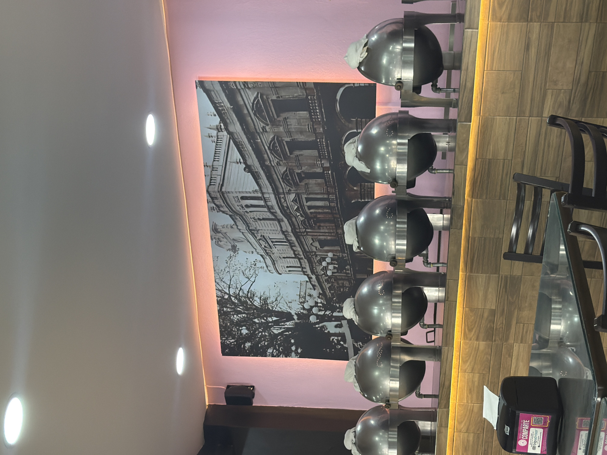 The image shows a restaurant interior with buffet warmers along a wall. A large black and white print of a building with tree branches is mounted on the wall above the buffet. The wall is light purple and has a warm light strip above. A table with a glass top and black chairs is visible. A black tissue box is on the table. The buffet warmers are silver and have white cloths inside. The location is Puebla, Mexico.