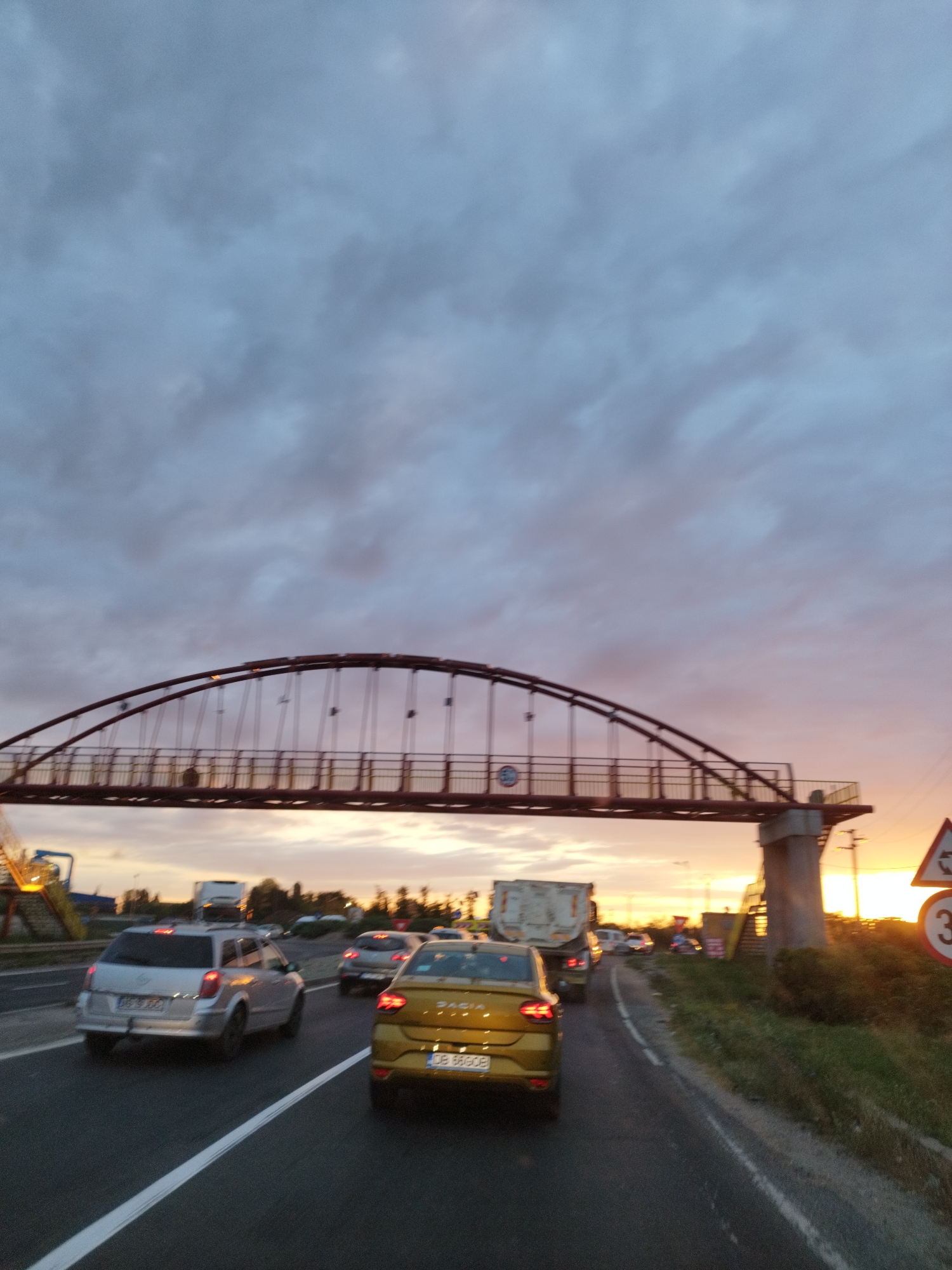 The image depicts a traffic scene on a road in Chitila, Romania, during what appears to be dusk or sunset. An arched pedestrian bridge spans the road, with its supporting structure visible on the right. Several vehicles are present, including a silver station wagon in the foreground, a gold sedan behind it, and various other cars and trucks further down the road, indicating a traffic jam or slow-moving traffic. The sky is cloudy with hues of orange and purple, characteristic of sunset. Road markings, including a white dashed line, are visible on the asphalt. Traffic signs, including a circular sign with the number "3" and a triangular sign, are also present on the right side of the road. The overall environment suggests a semi-urban or suburban setting.