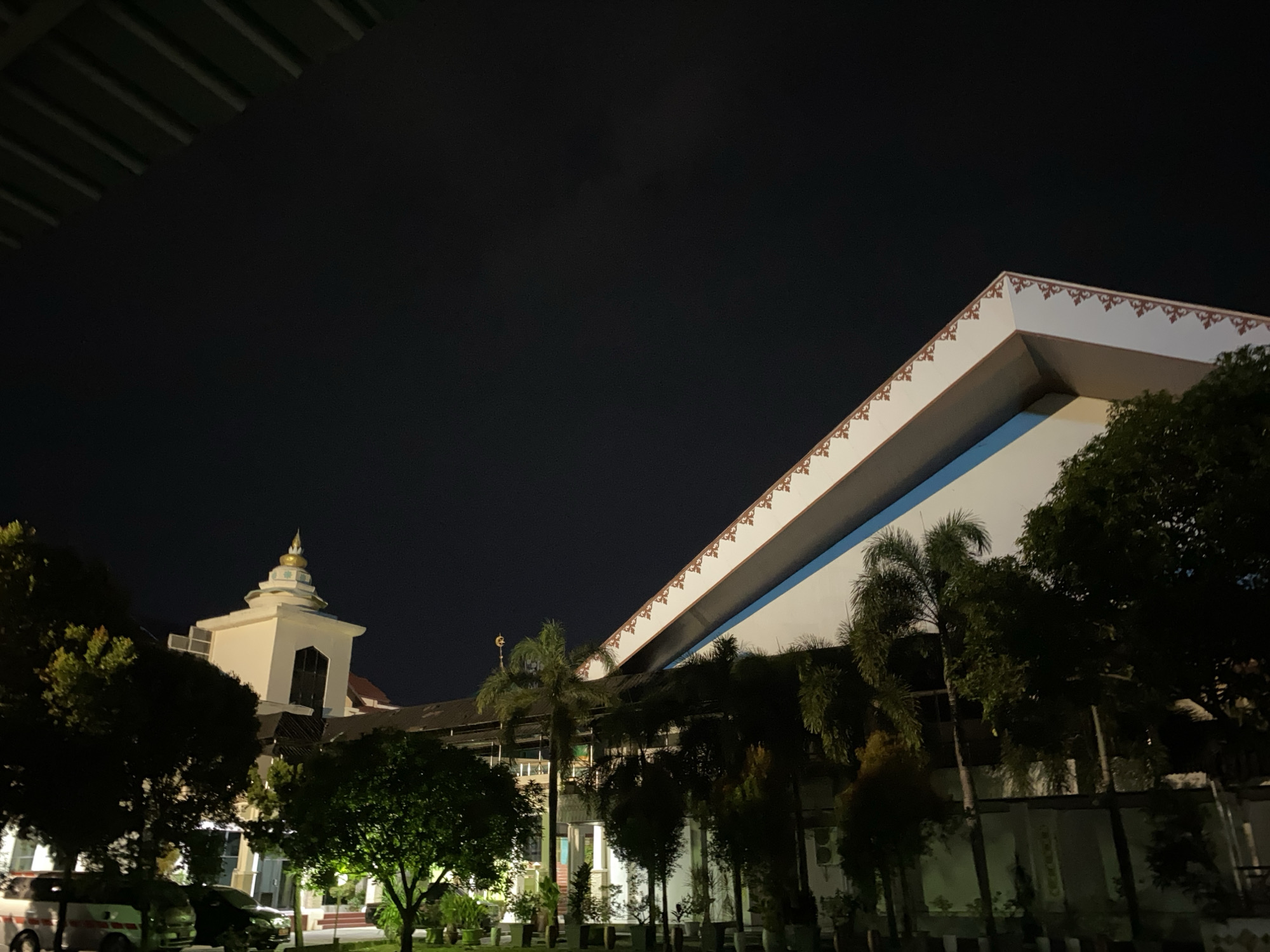 This image captures a building at night in Banda Aceh, Indonesia. The primary subject is a modern, multi-story building with a distinctive white facade and a sloped roofline adorned with a decorative border. The building is illuminated, highlighting its architectural features against the dark night sky.
To the left of the main building, a smaller, white structure with a domed roof and a golden spire is visible, suggesting a religious or cultural significance, possibly a mosque or a similar edifice. Several trees, including palm trees, are scattered in front of and around the buildings, with their foliage dark against the night. In the foreground, a few vehicles, including what appears to be a white van and a dark car, are parked on a paved area, indicating this might be a public space or a parking lot.
The scene is set at night, with the sky completely dark, devoid of stars or moon. The lighting on the building suggests it is an active location even after dark. The weather appears calm, with no signs of rain or wind. The overall mood is quiet and serene, typical of a nighttime scene in an urban or semi-urban environment. There is no visible text in the image.