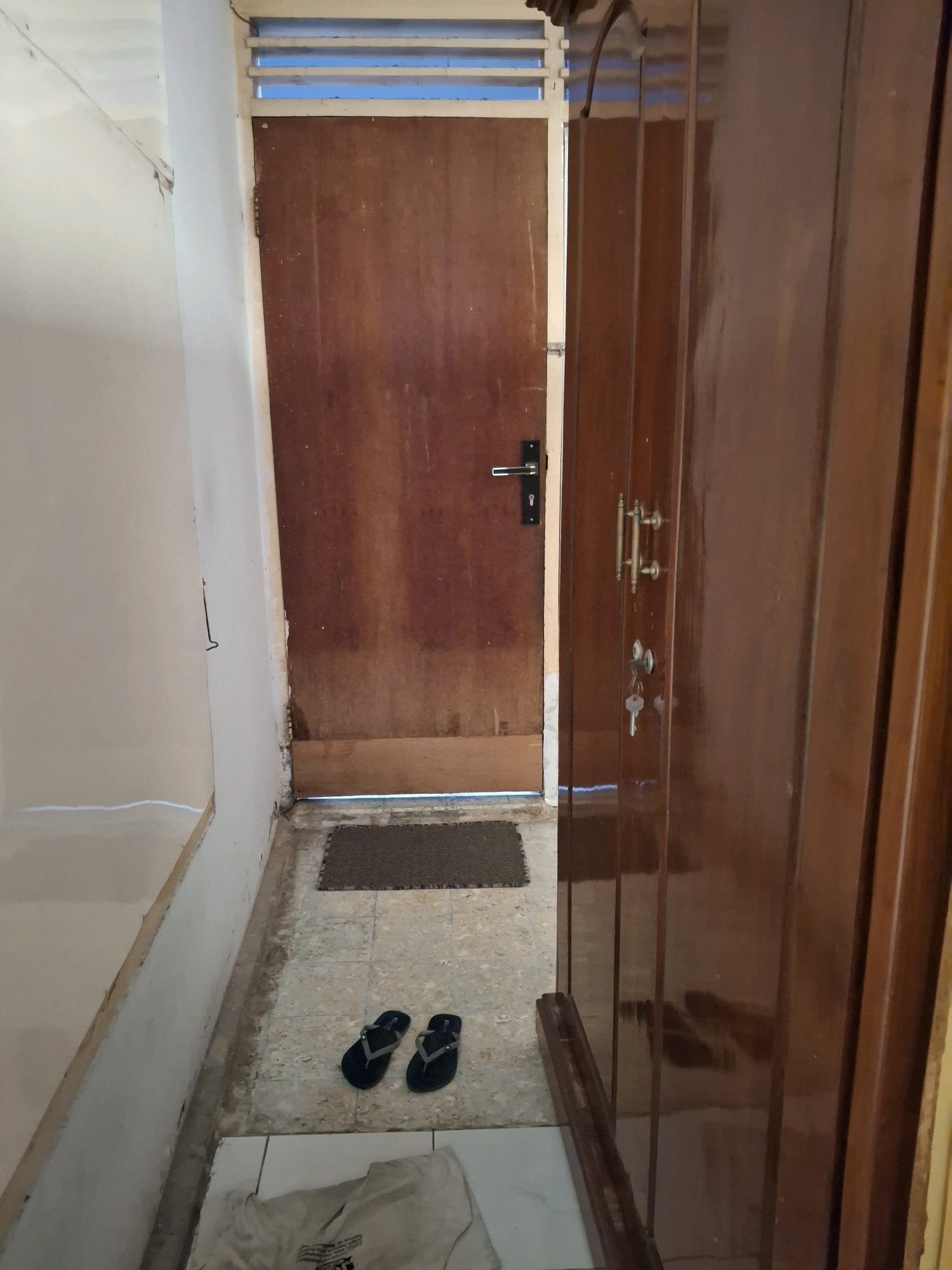 The image depicts an interior scene, likely a hallway or entryway, featuring two wooden doors. The door on the left is a reddish-brown, solid wood door with a silver lever handle and a visible keyhole. Above this door, a set of horizontal louvers is visible, suggesting a window or vent. The door on the right is a darker, polished wood with multiple panels and two decorative brass handles, one above the other. Below the upper brass handle, there are two keyholes, and keys are inserted into both. A second, similar door is partially visible to the far right.
On the floor in front of the doors, there is a small, dark doormat. To the left of the doormat, a pair of black flip-flops is laid out. A light-colored t-shirt or fabric item is lying on the floor in the foreground, partially obscuring the bottom edge of the image.
The walls are painted a light color, with one wall on the left appearing to be a textured or possibly damaged surface, showing some peeling or exposed material near the top. The floor is tiled with a light-colored, somewhat worn pattern. The lighting in the scene is somewhat dim.
The overall impression is of a functional, perhaps slightly dated, domestic interior. Without specific landmarks or signage, it is impossible to definitively place this scene in Lumajang, Indonesia, but the architectural style and materials are consistent with residential buildings found in Indonesia.