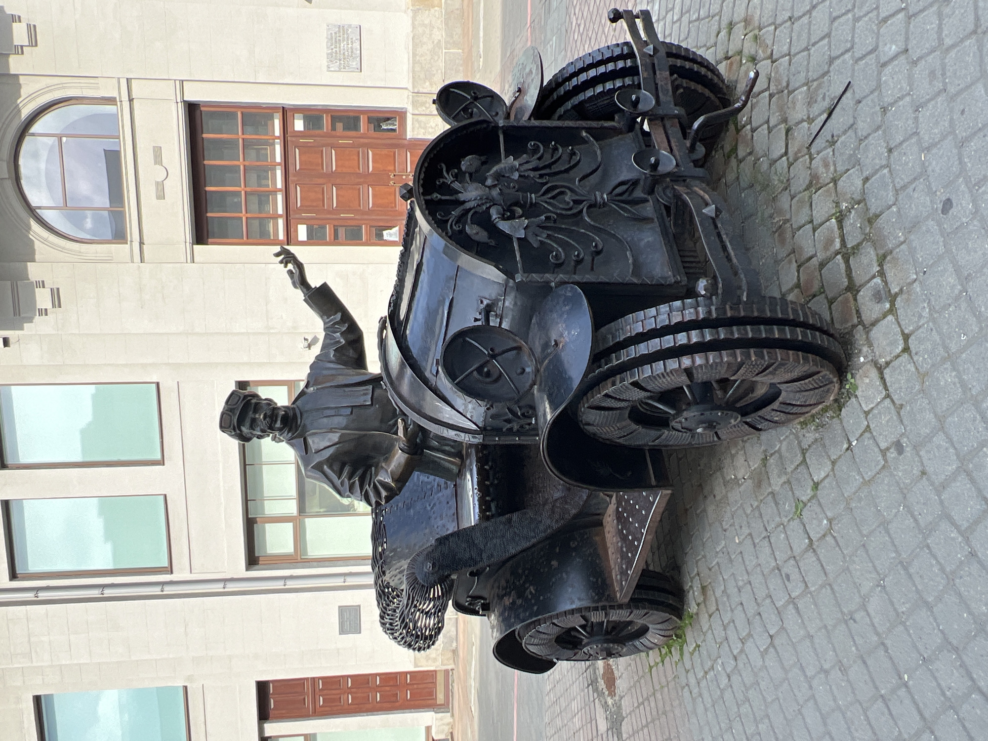 The image shows a bronze sculpture of a man in a hat and aviator goggles, sitting in a vintage-style car. The sculpture is located on a cobblestone street in front of a building with large windows and wooden doors. The man is depicted with his right arm extended and his hand open, as if speaking or gesturing. The car is elaborately decorated with wrought-iron details, including a floral design on the front grille. The sculpture is a monument to Ivan Ivanovich Ivlev, a pioneer of aviation and automobile engineering, and is situated in Yekaterinburg, Russia.