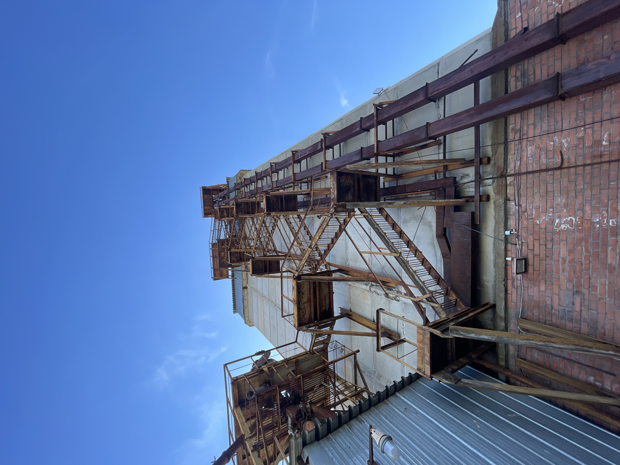 This media file captures the exterior of an aged industrial building against a clear, bright blue sky. The image is dominated by the building's raw, utilitarian architecture, characterized by extensive rust and weathered materials.
The central focus is a multi-story complex of rusty metal structures, including a series of steep stairs, platforms, and support beams that ascend the facade of the building. These metal elements exhibit significant corrosion, indicating prolonged exposure to the elements and age. The stairs have open risers and simple railings, appearing as a functional but well-worn access system, possibly a fire escape or maintenance access.
The building itself features at least two distinct wall types: a light-colored, possibly concrete or rendered masonry section that forms the main vertical structure, and a section of red brick visible on the right side of the frame. Heavy, rusty I-beams are horizontally integrated into the upper parts of both the concrete and brick walls, suggesting structural supports or former attachments for machinery.
In the lower foreground, a corrugated metal roof is partially visible, with a small light fixture attached to its edge. The sky, occupying a large portion of the upper left, is a vibrant, cloudless blue, indicating clear weather conditions during the daytime.
The scene conveys a sense of industrial decay and history, typical of an old factory, silo, or other heavy industry infrastructure. No human activity or visible text is present. The lighting is natural daylight, highlighting the textures and rust of the metal and brickwork. The location context for this industrial scene is Krasnoyarsk, Russia.