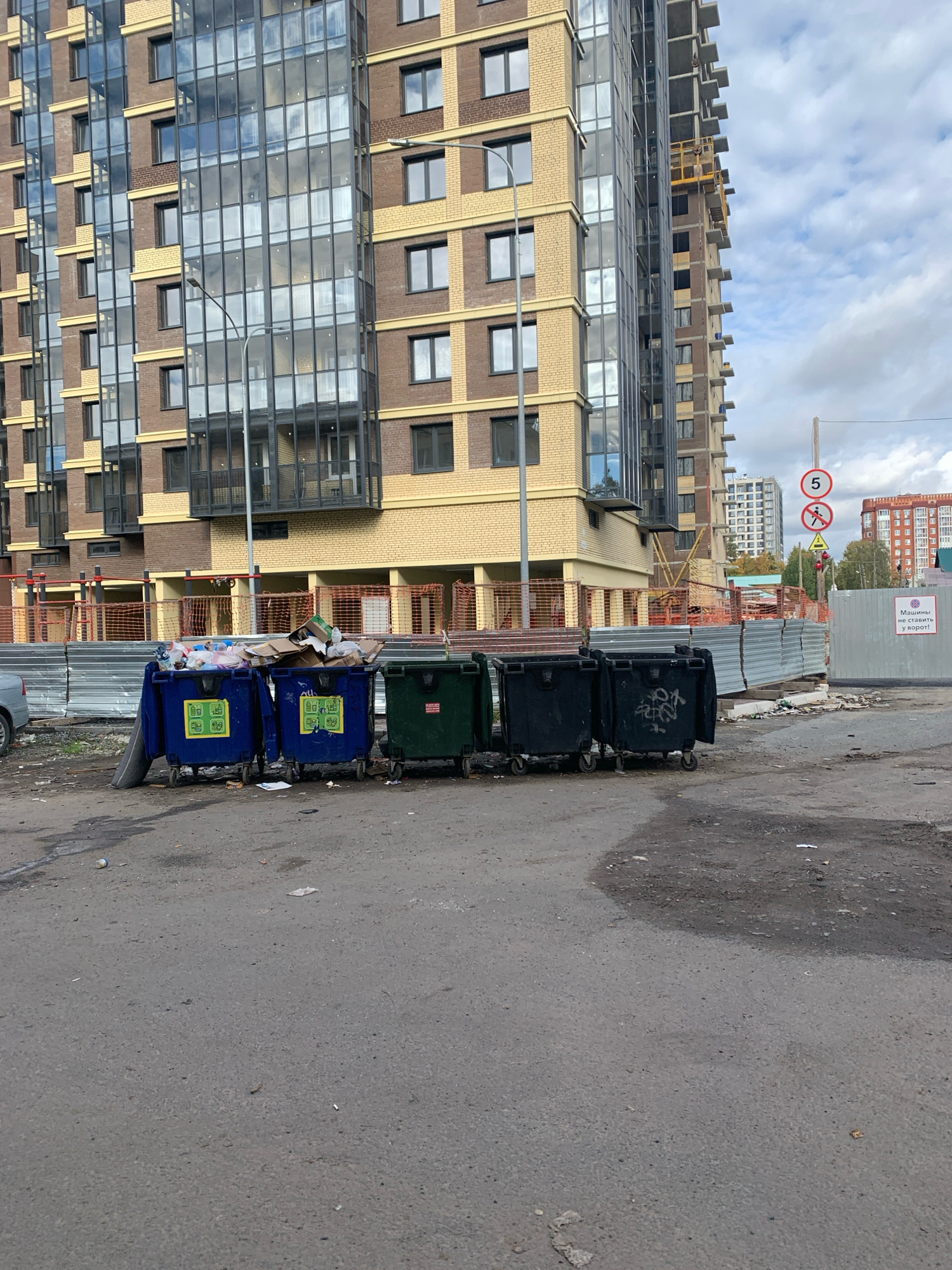 The image depicts a construction site in Arkhangelsk, Russia, featuring a partially constructed multi-story building with a facade of brick and glass. In the foreground, several large, wheeled garbage bins are filled with refuse. Two blue bins are prominently displayed, filled with a variety of trash, including cardboard boxes, plastic bags, and other debris. To their right are two dark green or black bins, also containing trash. The ground is a paved surface, appearing somewhat worn and littered with smaller pieces of debris. A temporary fence made of corrugated metal sheets is visible behind the bins, and orange construction netting is strung across a section of the site. In the background, other buildings, including what appears to be another apartment complex, can be seen. Traffic signs, including a speed limit sign (5) and a prohibition sign, are also present. The sky is overcast, suggesting a cloudy day. No people are visible in the image.