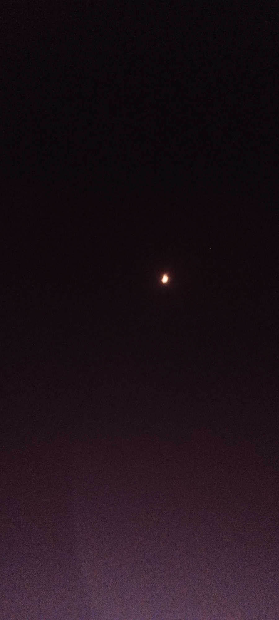 The image shows a dark night sky with a crescent moon glowing in the upper center. The moon appears to be partially illuminated and has a reddish-orange hue. There is a small, faint point of light to the right of the moon, possibly another celestial body. The lower portion of the image displays a gradient of dark purple and pink, suggesting a low horizon or atmospheric conditions. There are no people, objects, or actions visible in the scene. The scene depicts a clear night sky over what is presumed to be the city of Adai, Spain, characterized by the presence of celestial bodies.