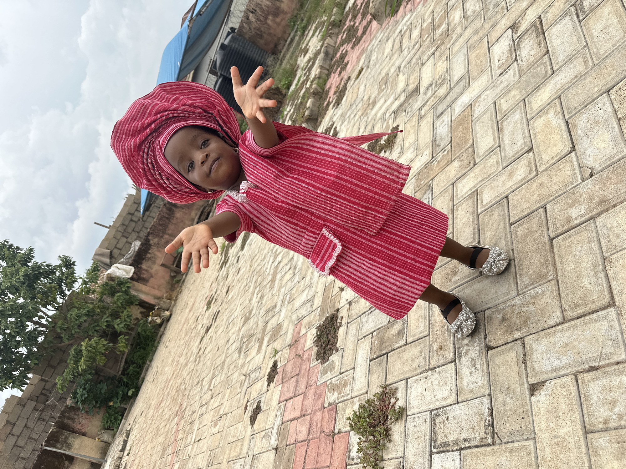 A young Nigerian girl, likely under five years old, stands on a paved courtyard in Jos, Nigeria. She is adorned in a traditional red and white striped outfit and a matching headwrap. Her arms are outstretched, palms up, as if offering a greeting or inviting play. She wears sparkling silver ballet flats. The courtyard's paving stones are arranged in a herringbone pattern. In the background, a weathered brick wall, overgrown with vines, suggests an older structure. Beyond the wall, a glimpse of a blue tarped roof and water tanks indicates a residential or commercial area. The sky above is overcast with scattered clouds, typical of the Nigerian climate. The scene captures a moment of childlike innocence against an urban backdrop.