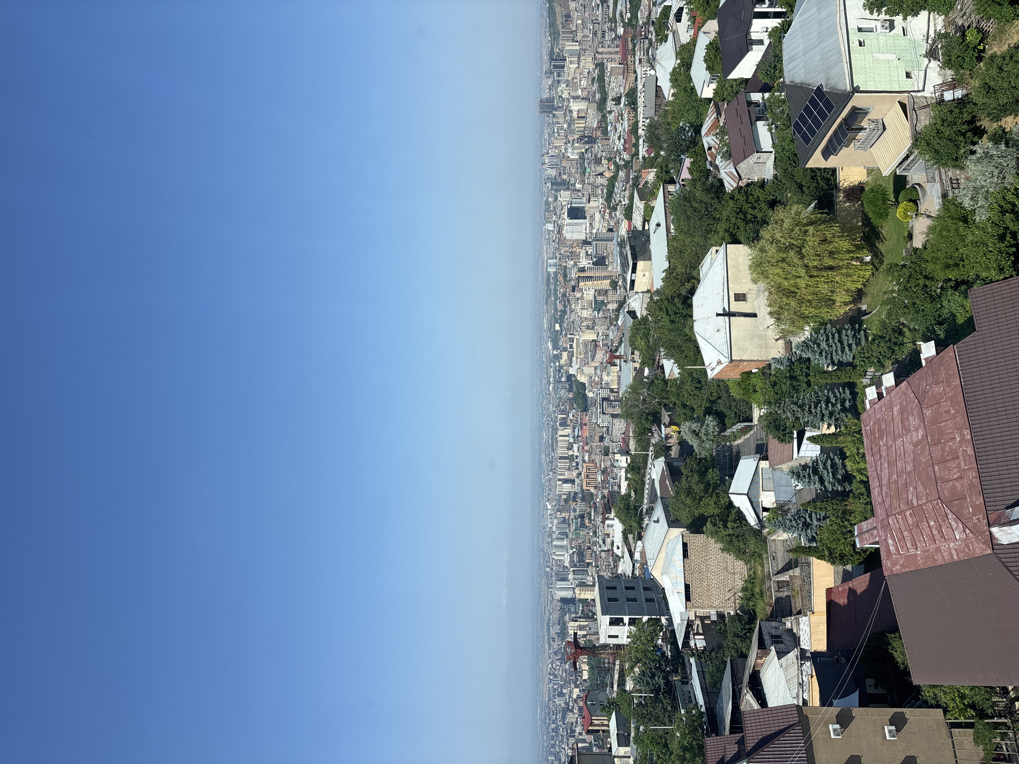 An aerial view captures a sprawling cityscape of Yerevan, Armenia, under a clear, bright blue sky. The foreground is dominated by the rooftops of residential buildings, characterized by diverse roofing materials such as brown and red corrugated metal, and some lighter-colored, sloped roofs. Dense green foliage from trees is interspersed throughout this area, partially obscuring some of the structures.
Further into the midground, the urban landscape expands, showcasing a mix of apartment buildings and smaller houses. Distinctive architectural features include a modern, multi-story building with a white facade and numerous windows, and another structure with a rough, stone exterior. A red construction crane is visible in the distance, indicating ongoing development.
The background reveals the extensive expanse of Yerevan, with a dense concentration of buildings stretching towards the horizon. Many taller, modern buildings with flat roofs are present, interspersed with older structures. The distant landscape is hazy, suggesting a wide urban area. The overall impression is of a vibrant, densely populated city on a sunny day.