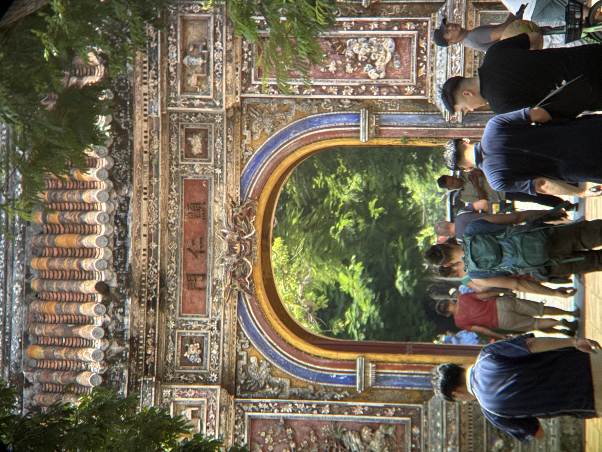 The image depicts the Ngo Mon (Meridian Gate) in the Imperial City of Huế, Phú Xuân Ward, Vietnam. A group of people are passing through the ornate archway. In the foreground, a man in a blue polo shirt with a yellow collar is walking away from the camera, with his back to the viewer. To his right, another man in a black t-shirt is also walking away. Behind them, a man with a backpack walks through the arch. Other individuals are visible further back, some appearing to be tourists. The gate itself is elaborately decorated with intricate carvings, colorful tiles on the roof, and architectural details characteristic of Vietnamese imperial structures. Lush green trees are visible through the archway, indicating an outdoor setting. The overall impression is of a historical landmark bustling with activity.