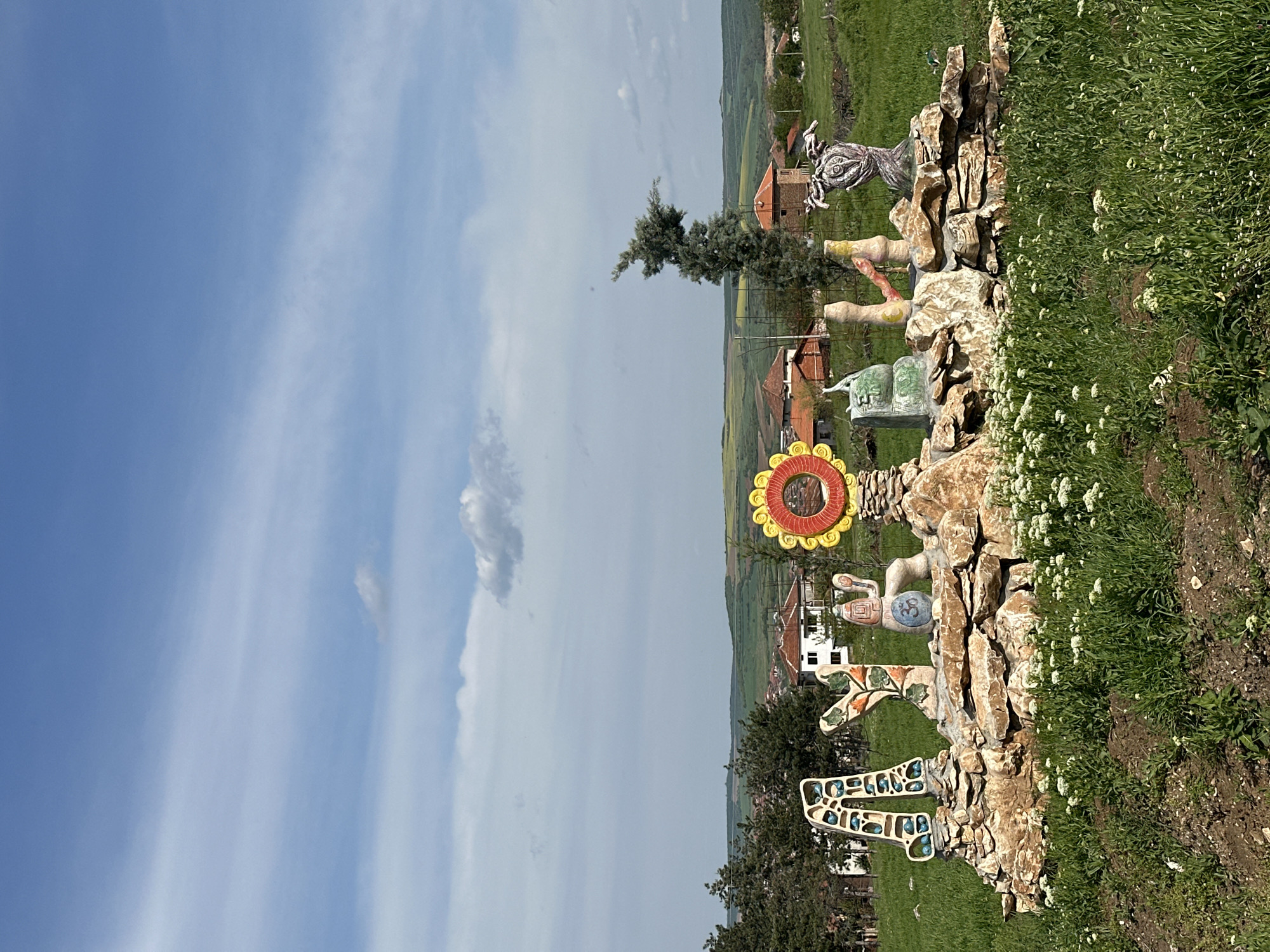 The image depicts an outdoor art installation in Lukovit, Bulgaria. Constructed along a low rock wall is a series of concrete sculptures, including a Medusa-like figure, a human-like torso, a turtle with the number "33," a stylized sunflower, a giraffe, and an abstract geometric design. These sculptures are situated in front of a green field with scattered buildings in the background under a blue and cloudy sky. The installation is partially bordered by a dense patch of white wildflowers and various types of vegetation. The image is oriented vertically with the art installations on the right side.