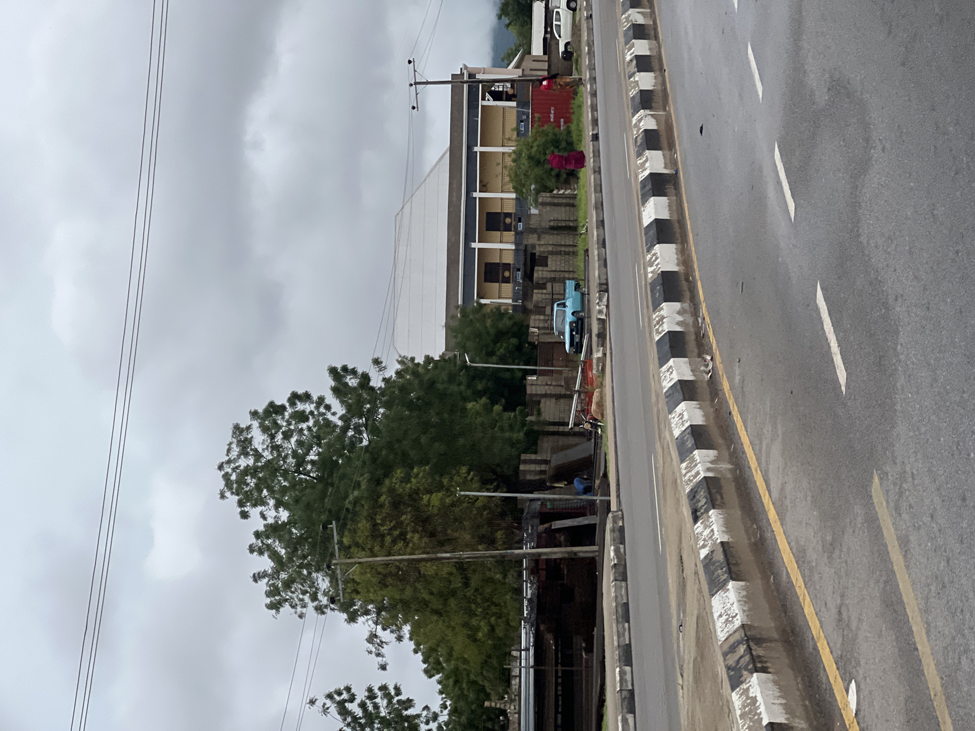 This is an exterior, eye-level, medium shot of a street scene in Jalingo, Nigeria, on an overcast day. In the foreground, a paved road with a yellow line and white dashed lines is visible, bordered by a concrete curb painted with alternating black and white blocks. Behind the curb, a strip of green grass and dirt separates the road from a building and trees.
The building in the midground is a two-story structure with a light-colored, sloped roof. The upper level features a yellow facade with dark windows and a covered balcony supported by white columns. To the left of the building, a light blue pickup truck is parked on a patch of ground. Several stacks of concrete blocks and metal bars are visible in front of the building and to its left. Two individuals are standing in front of the building; one, wearing a red garment, stands near a large red container, and the other, in a lighter-colored outfit, stands further to the right.
Behind the building and stretching across the midground are numerous mature trees with dense green foliage. A wooden utility pole with visible wires stands near the left side of the image, among the trees. Further in the background, more trees and a light-colored vehicle are partially visible. The sky is heavily overcast, suggesting cloudy or potentially rainy weather.