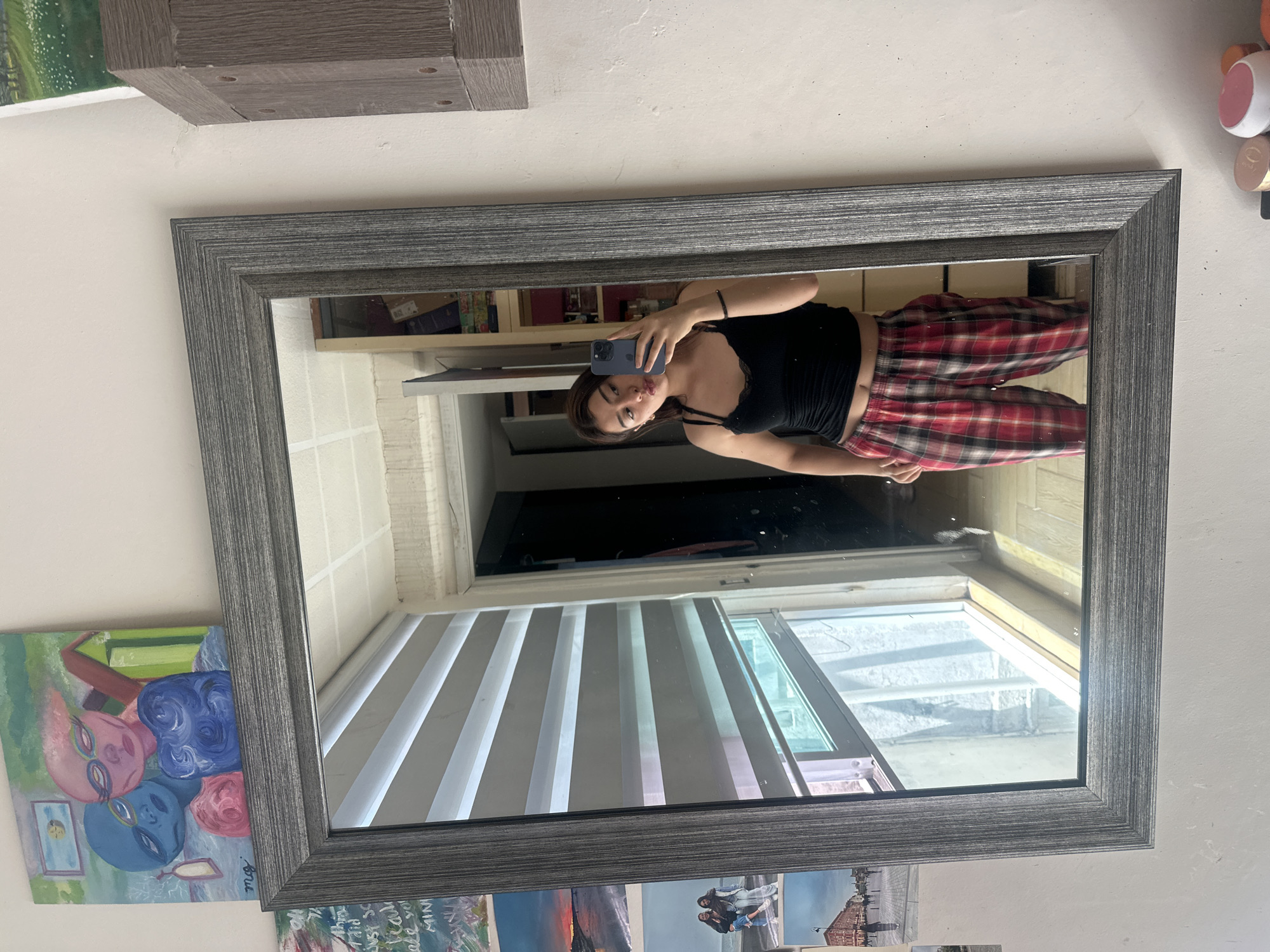 A woman stands in front of a mirror, taking a selfie with her smartphone. She is wearing a black tank top and plaid pajama pants. The mirror reflects a room with blinds on a window. The room appears to be an apartment, decorated with artworks. Given the decor in the reflection and the presence of artwork and other decorative items, it is difficult to establish if this is the city of Puebla in Mexico.